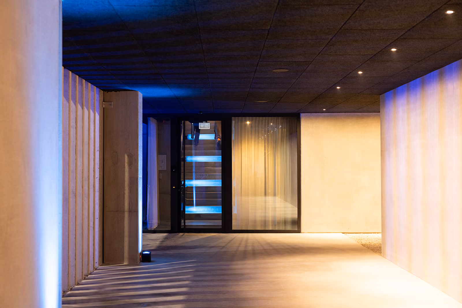 Moderne entreehal verlicht met blauwe en gele verlichting, glazen deur en zicht op trap binnenin.