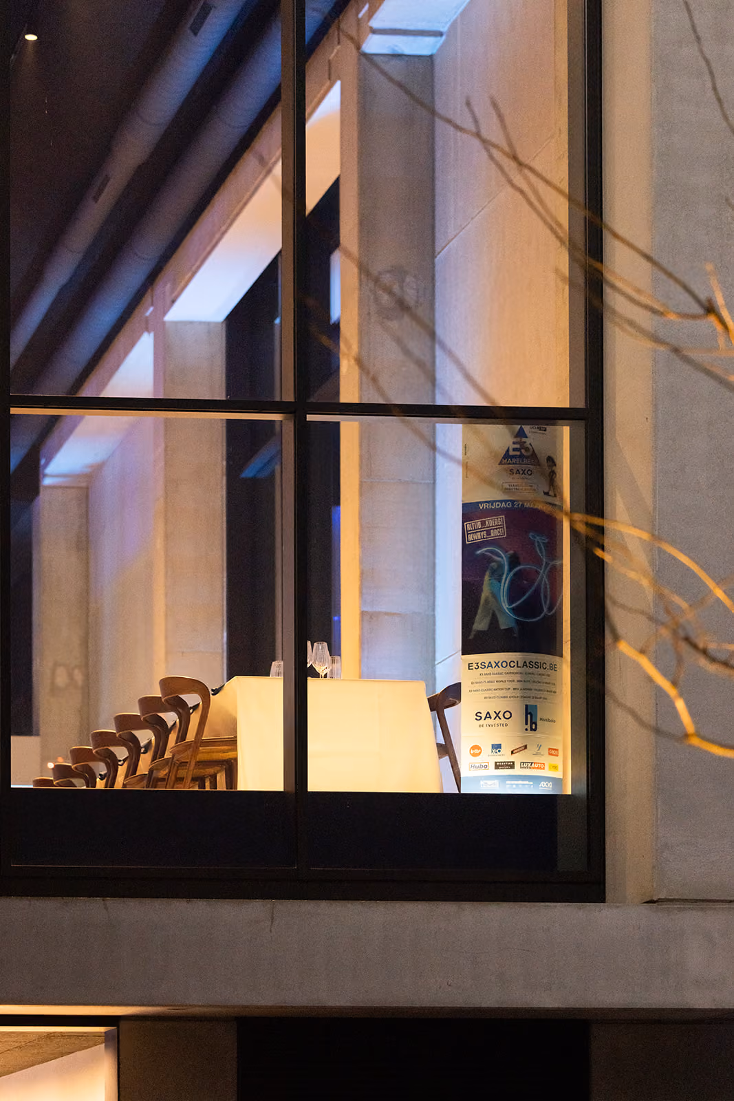 Gezette tafel met glazen en stoelen achter groot raam in modern gebouw bij avondlicht.