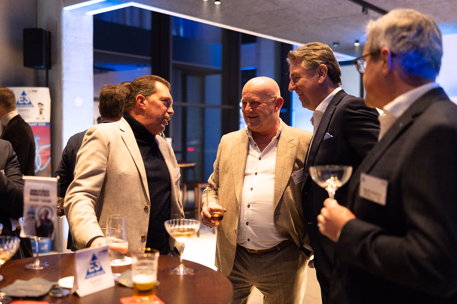 Vier mannen in elegante jassen praten en lachen bij een tafel met drankjes op een evenement.