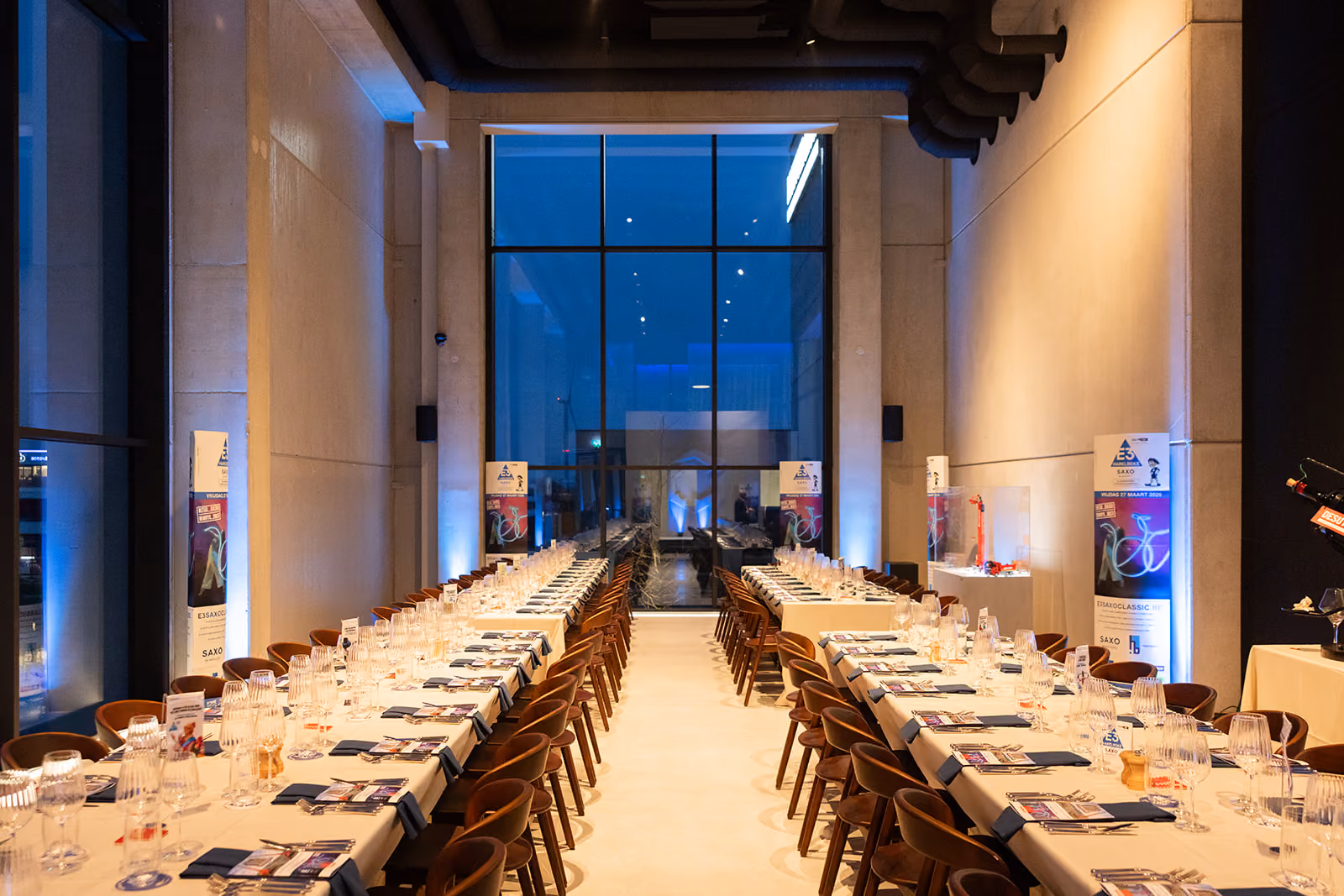 Lang gedekte tafels in een moderne zaal met glazen wand en warme verlichting.