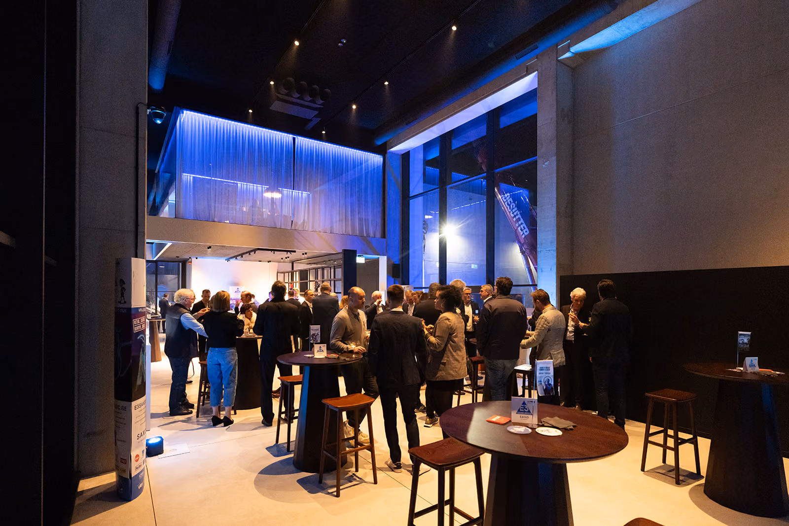 Mensen praten en drinken aan sta-tafels in een moderne, verlichte zaal met grote ramen.