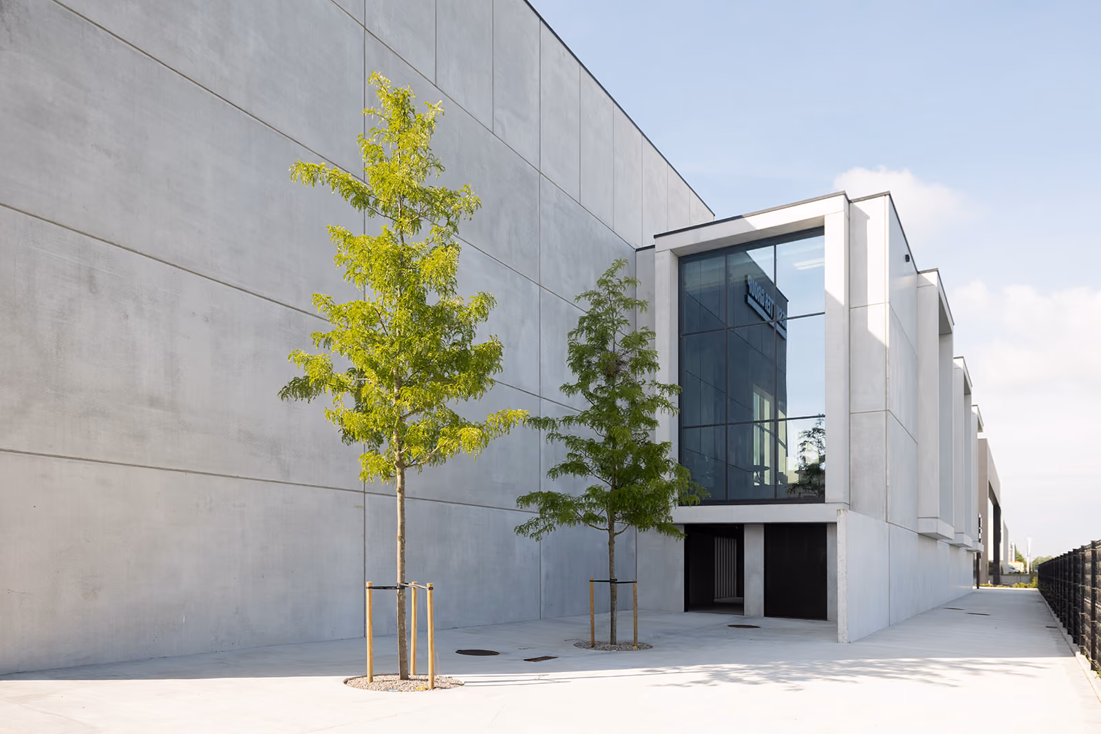 Moderne betonnen industrieel gebouw met twee jonge bomen en grote glazen ramen.
