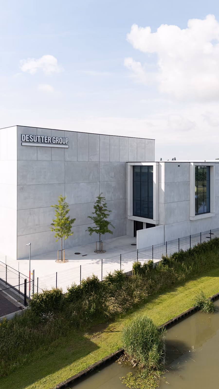 Modern concrete building with DESUTTER GROUP sign, two trees outside, and canal with grass in front.