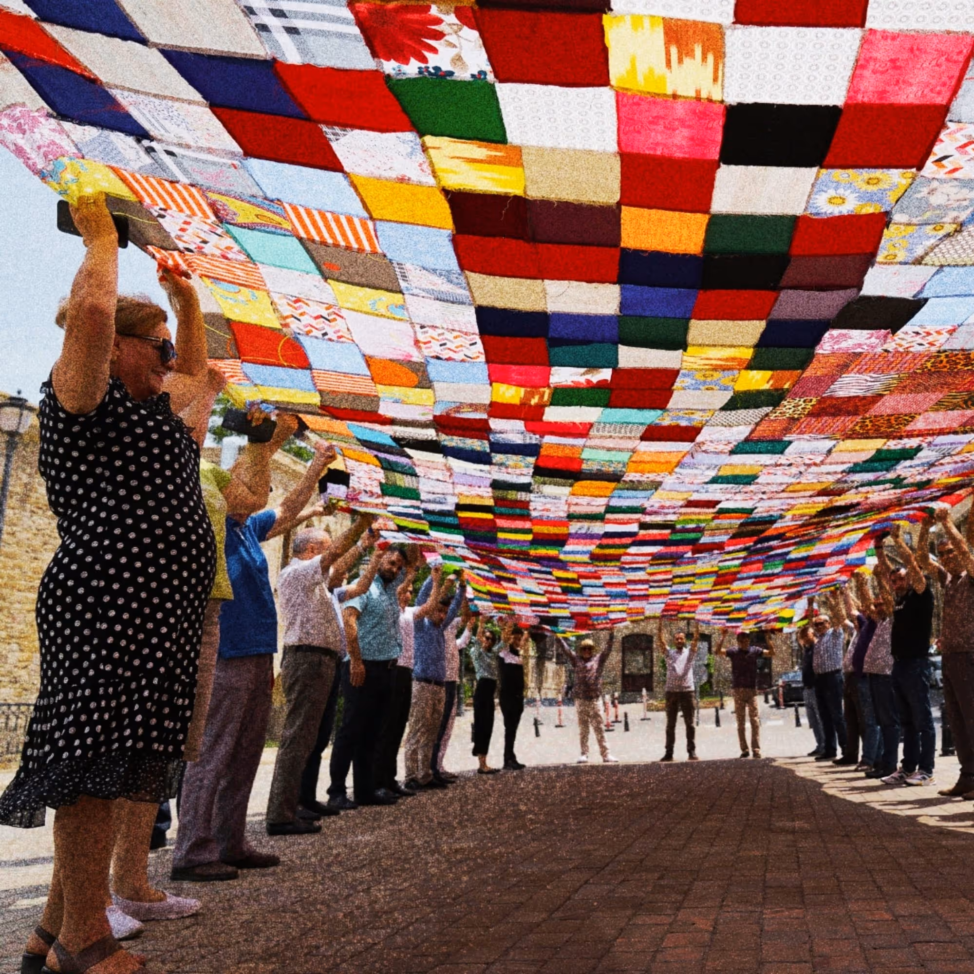 Group of people standing in a circle outdoors, raising a large colorful patchwork quilt above their heads.