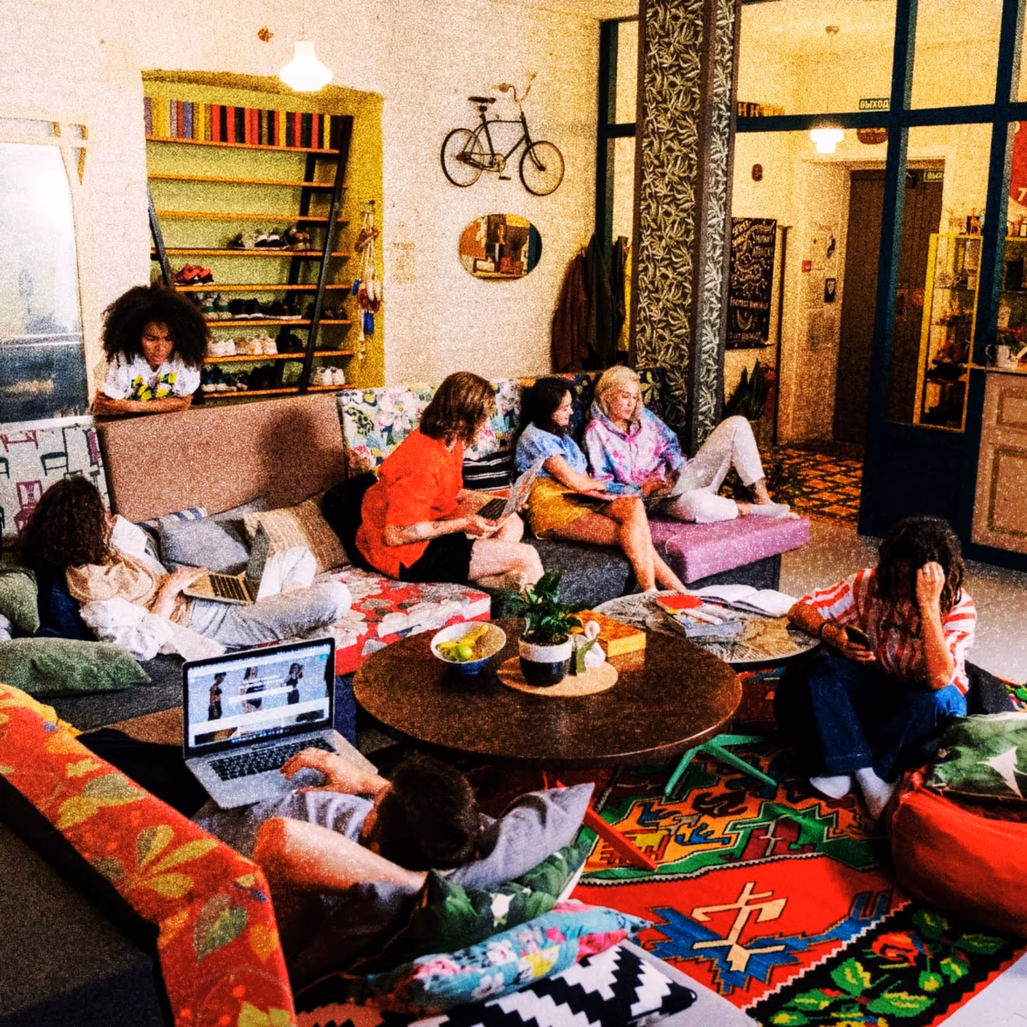 A colorful co-living space with seven people sitting on couches and floor cushions using laptops and phones.
