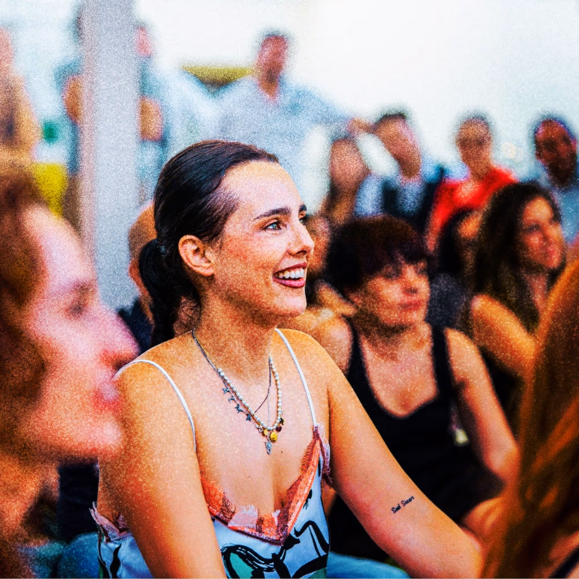 Smiling woman with dark hair tied back wearing a sleeveless top, seated among a group of people in a lively social setting.