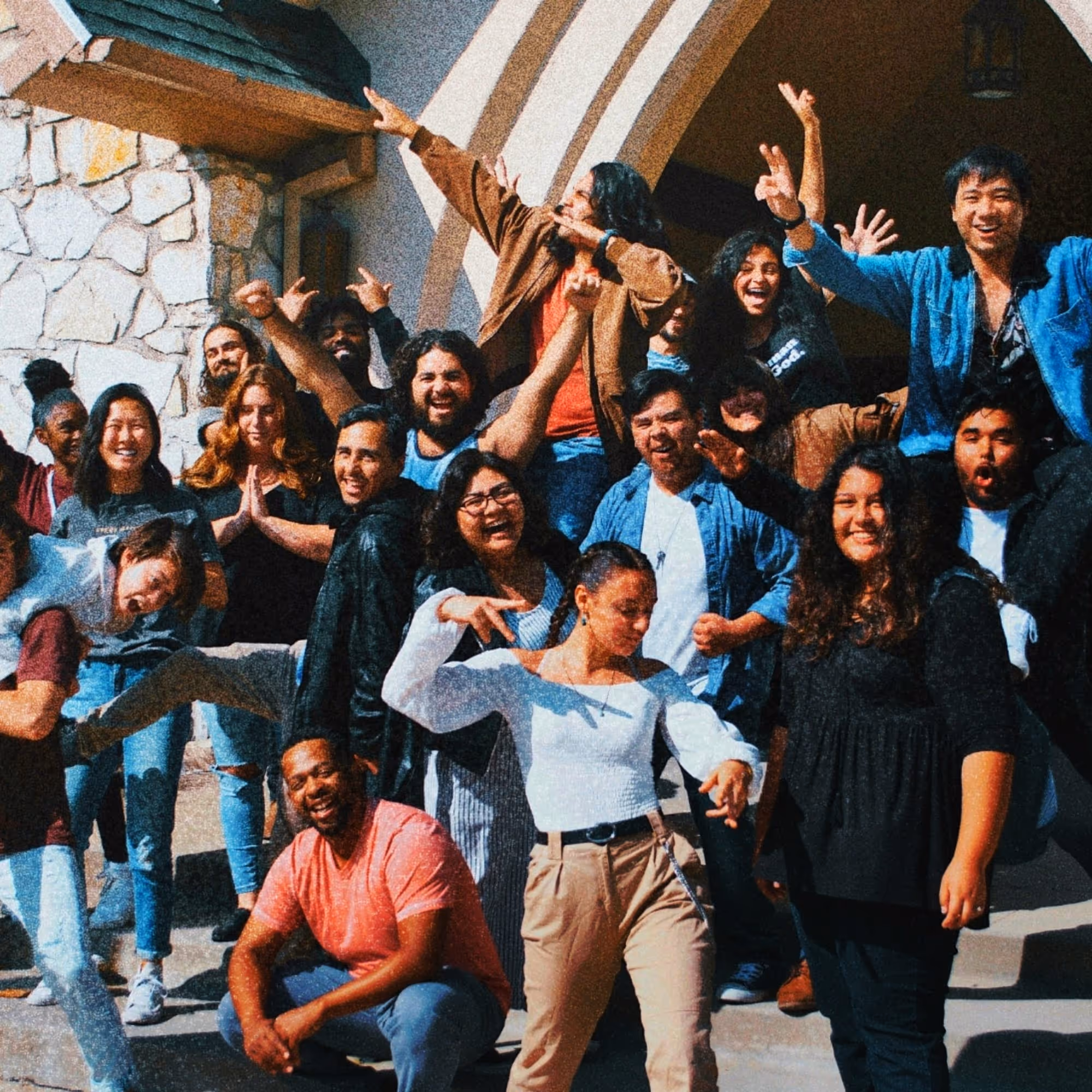 Group of diverse young adults smiling, posing, and making energetic gestures outside a stone building with an arched entrance.