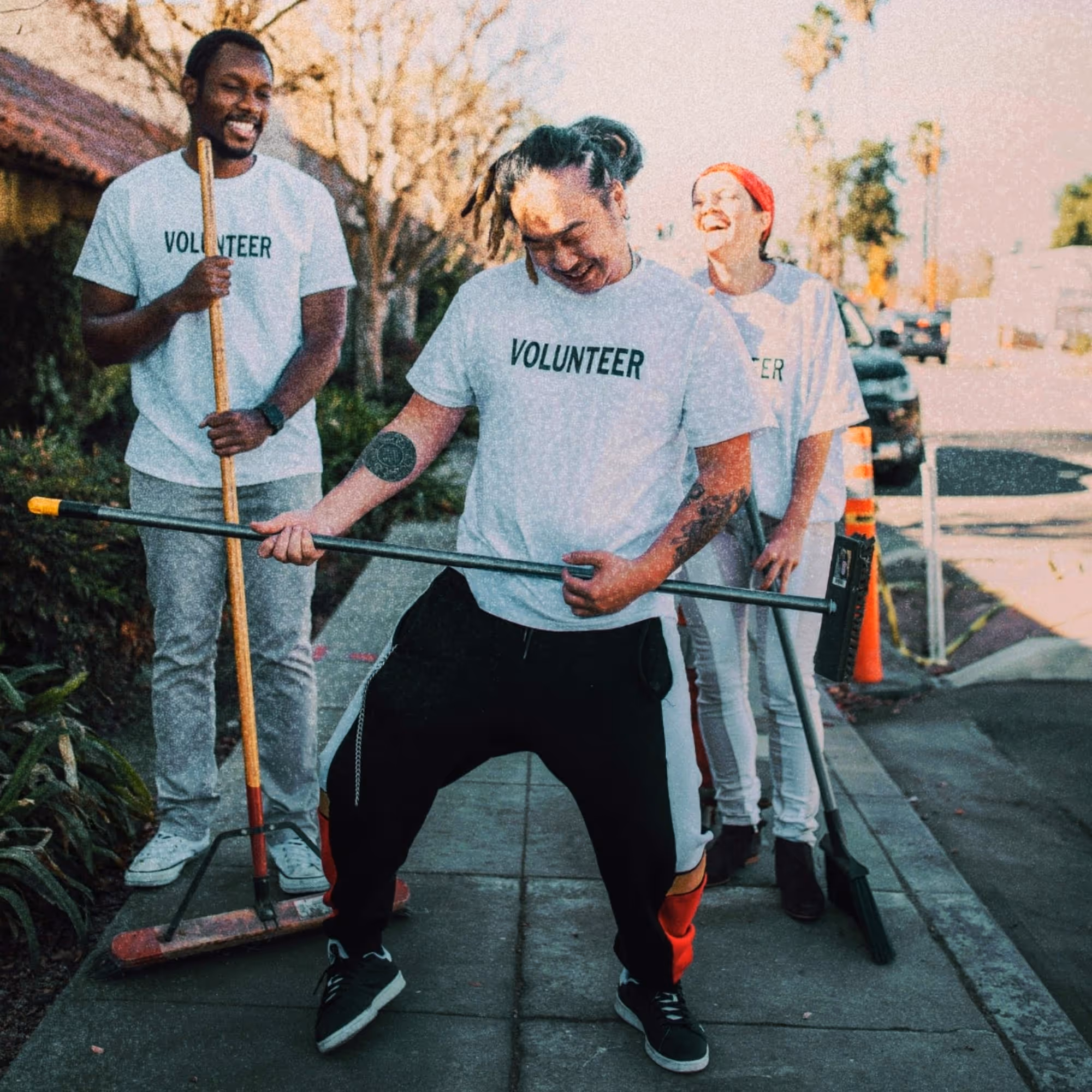 Three volunteers on a sidewalk, one pretending to play a broom like a guitar while the others laugh.