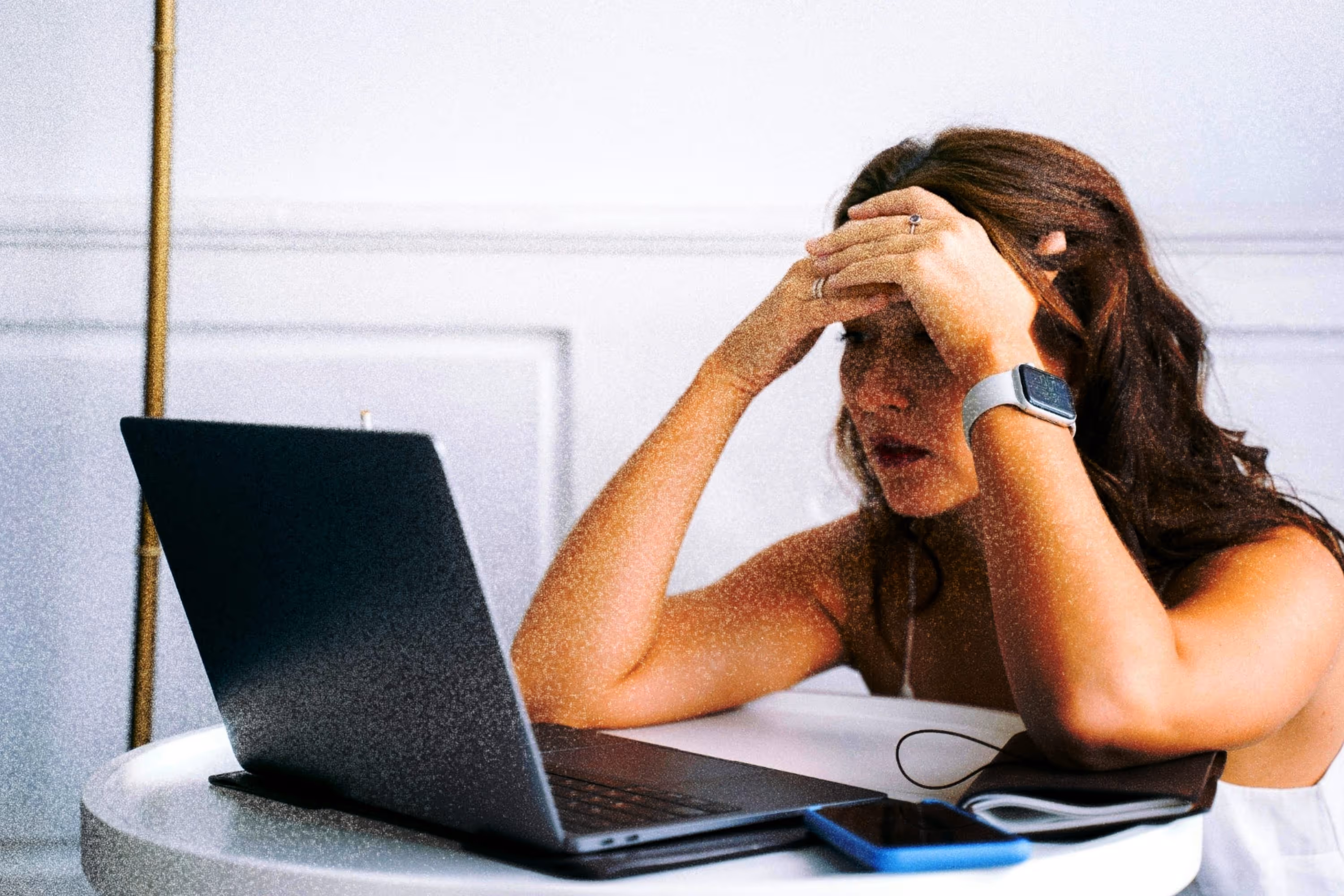 Woman sitting at a round white table holding her forehead, looking stressed while facing an open laptop with a smartphone beside it.