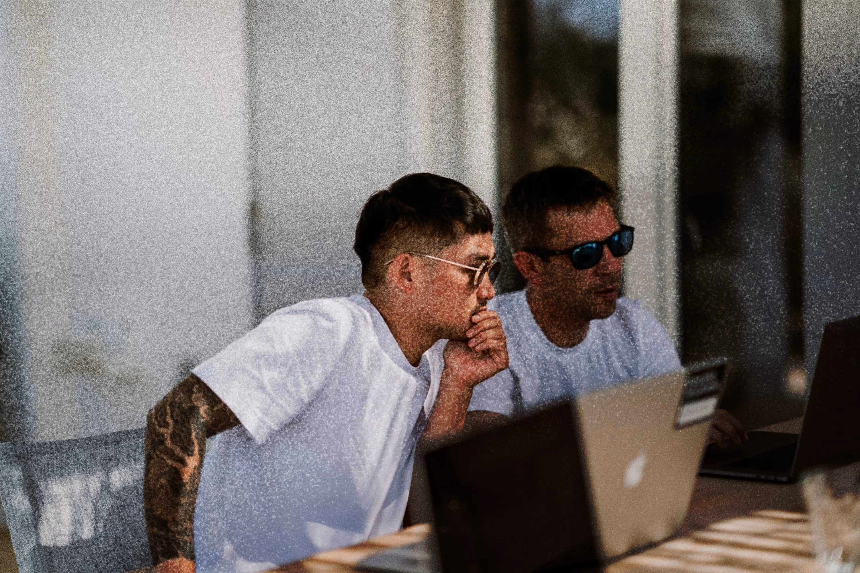 Two men wearing white t-shirts and sunglasses looking intently at laptop screens while sitting at a table.