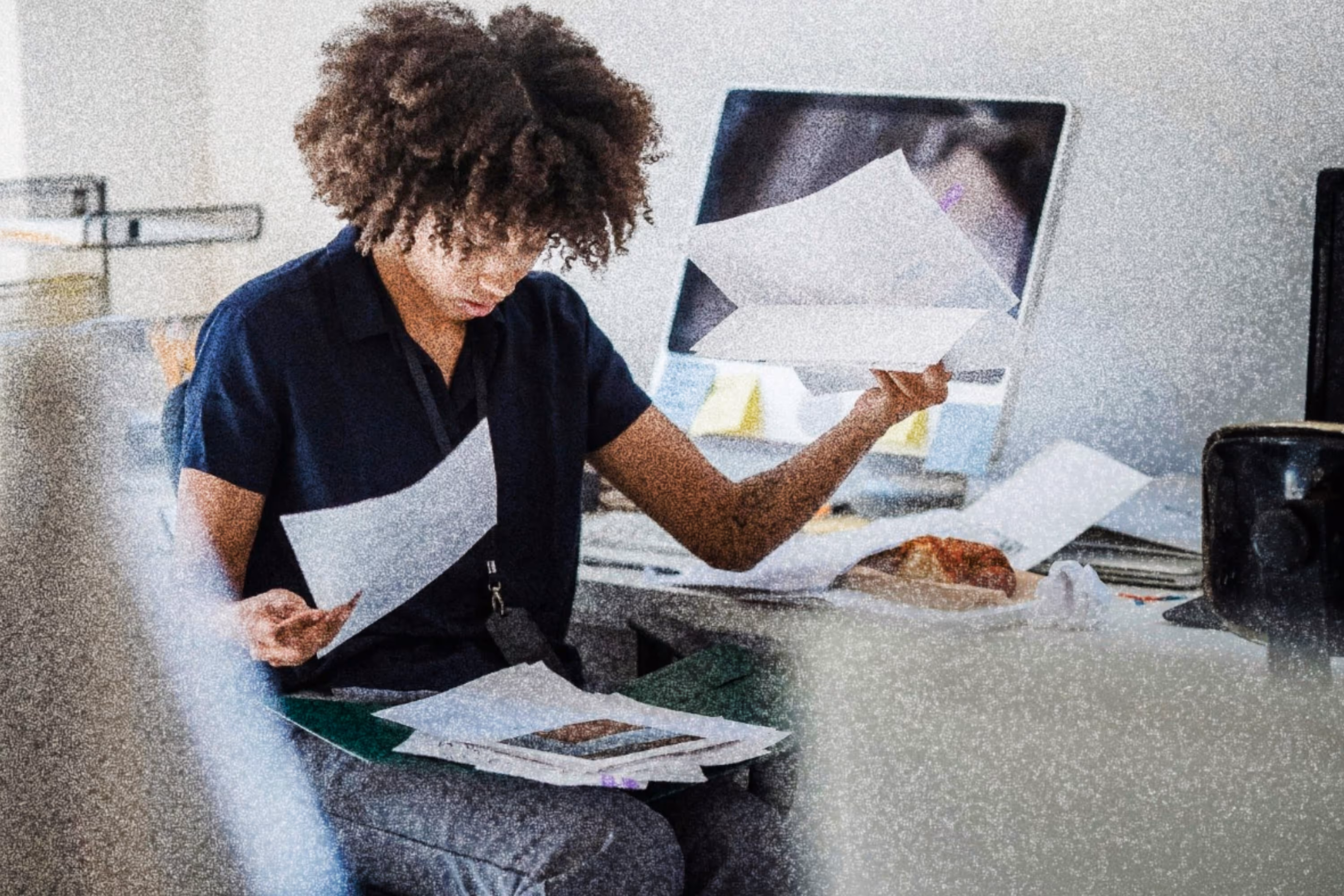 Woman sitting at a desk with scattered papers, looking focused while holding documents in both hands.