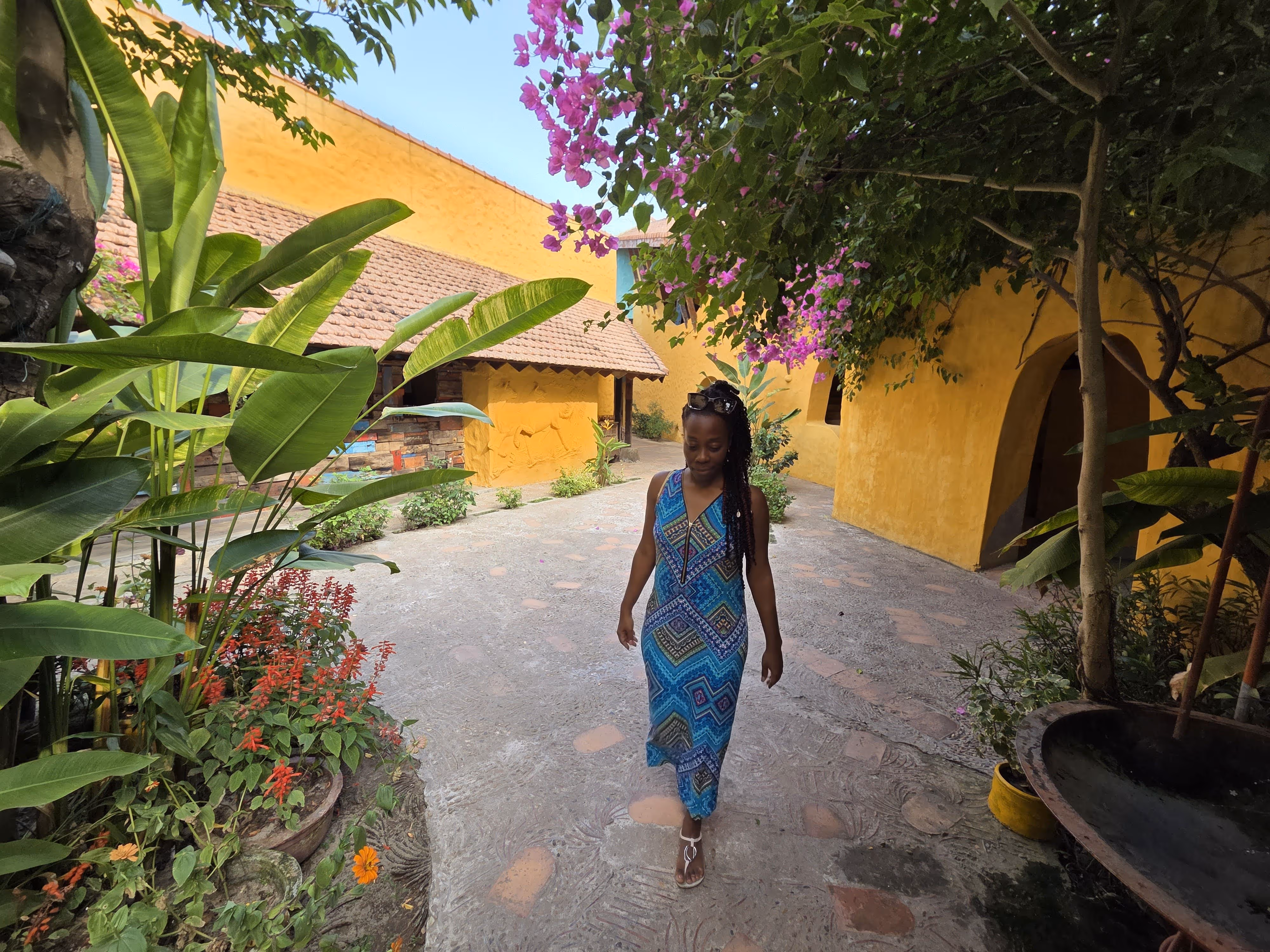 Woman in a blue patterned dress walking on a stone path surrounded by green plants and yellow walls with a tiled roof.
