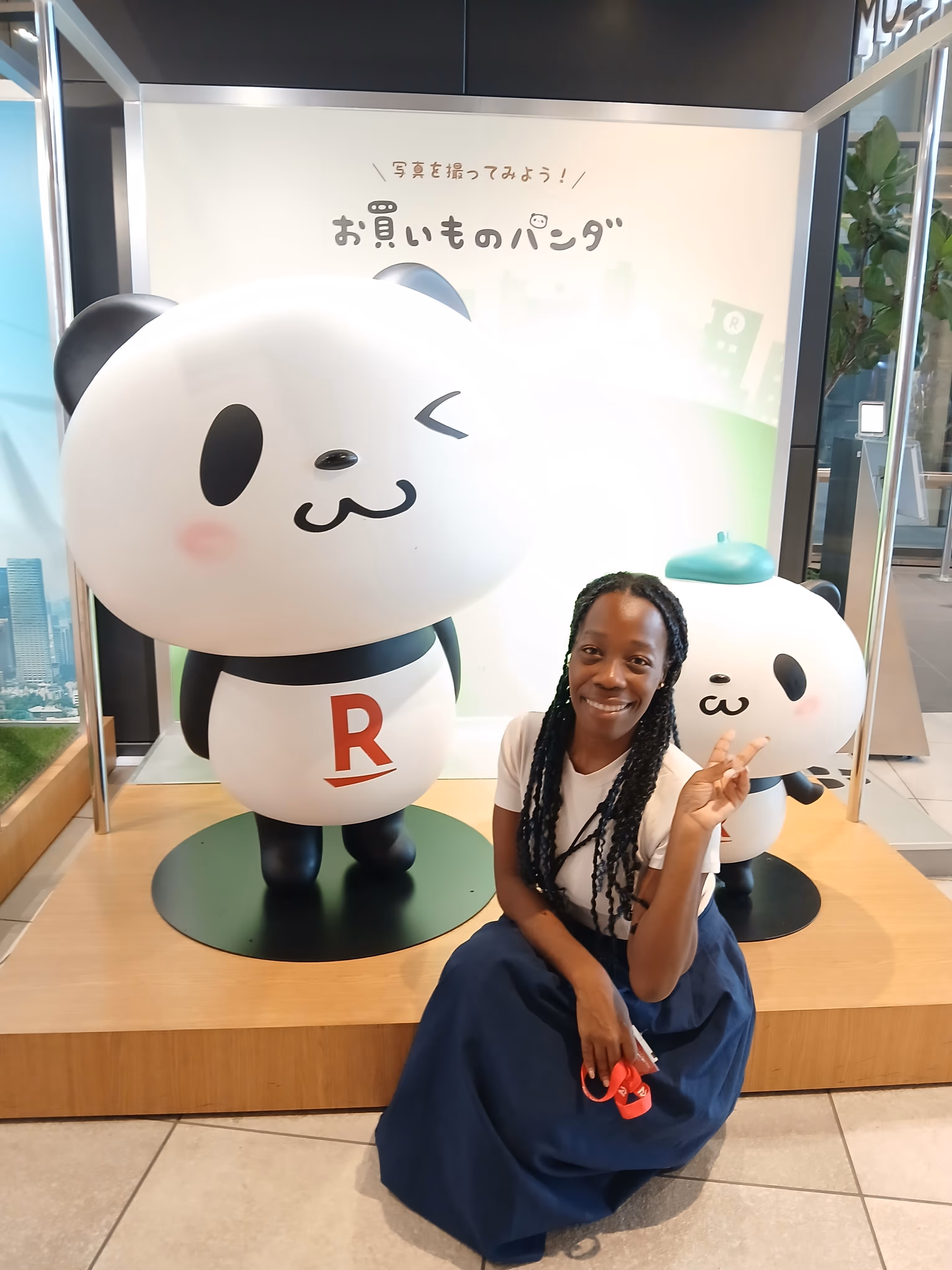 Smiling woman sitting on the floor next to two large winking panda mascots, one wearing a green beret, in a brightly lit indoor display area.