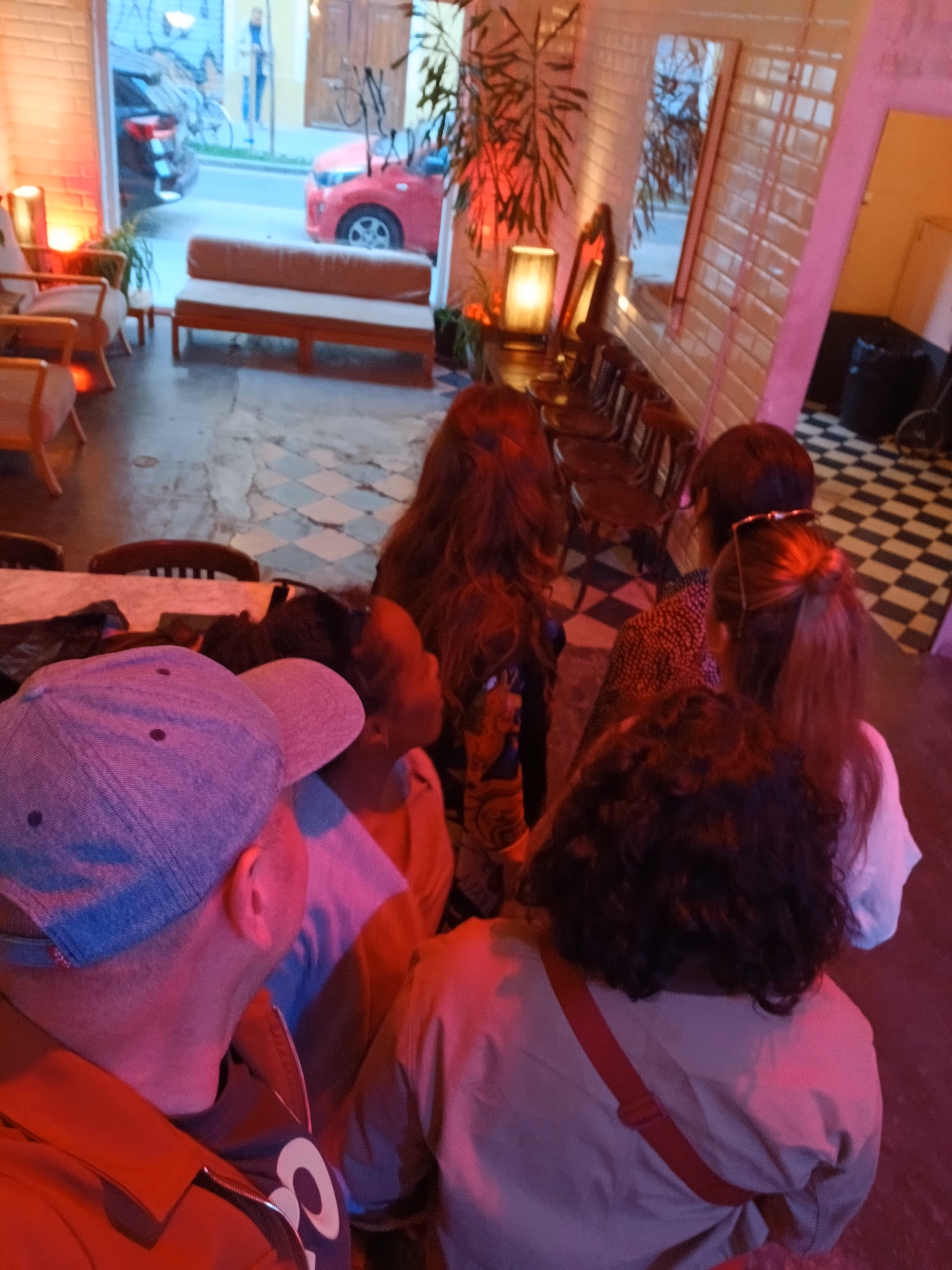Group of people standing indoors facing a large window with parked cars outside in a dimly lit room with checkered floor tiles and seating.