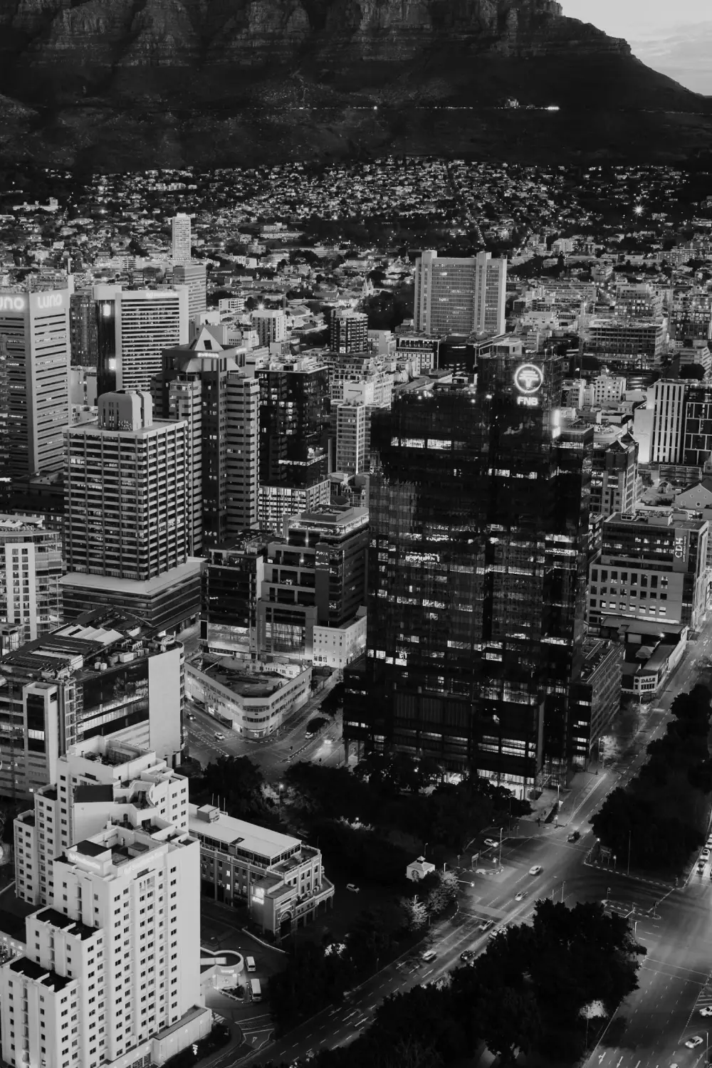Aerial view of a cityscape at dusk showing high-rise buildings, illuminated streets, and a mountainous backdrop.