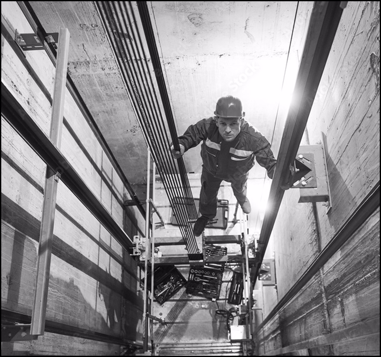 Technician in a hard hat and work uniform inspecting elevator cables inside a maintenance shaft.