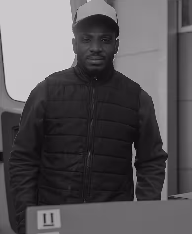 A man wearing a cap and jacket, wheeling in a package from a van.