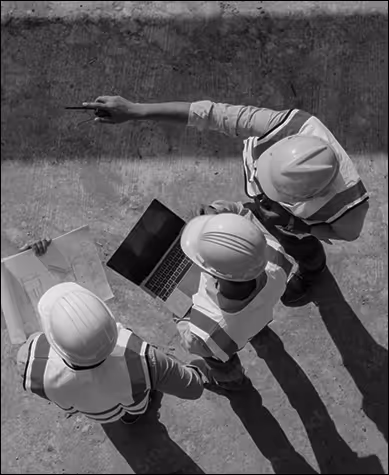A top down view of three standing people looking at a laptop and papers, and one of them are pointing out in a direction.