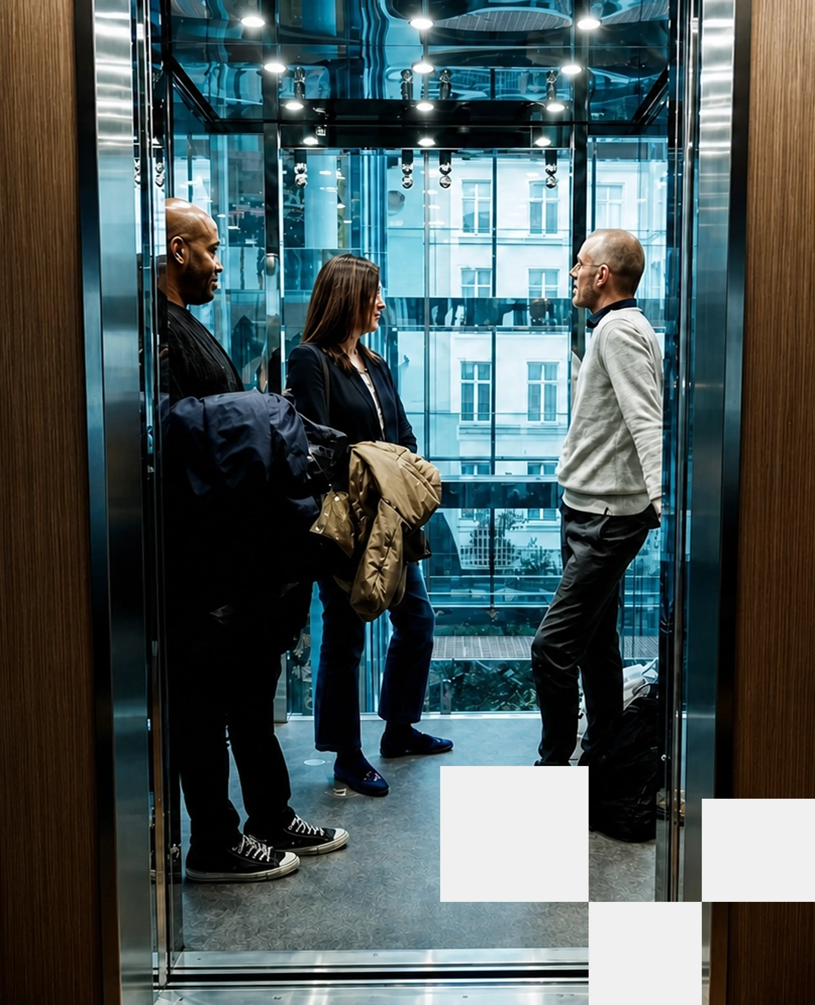 Three people standing and conversing inside a modern glass elevator with a cityscape visible outside.