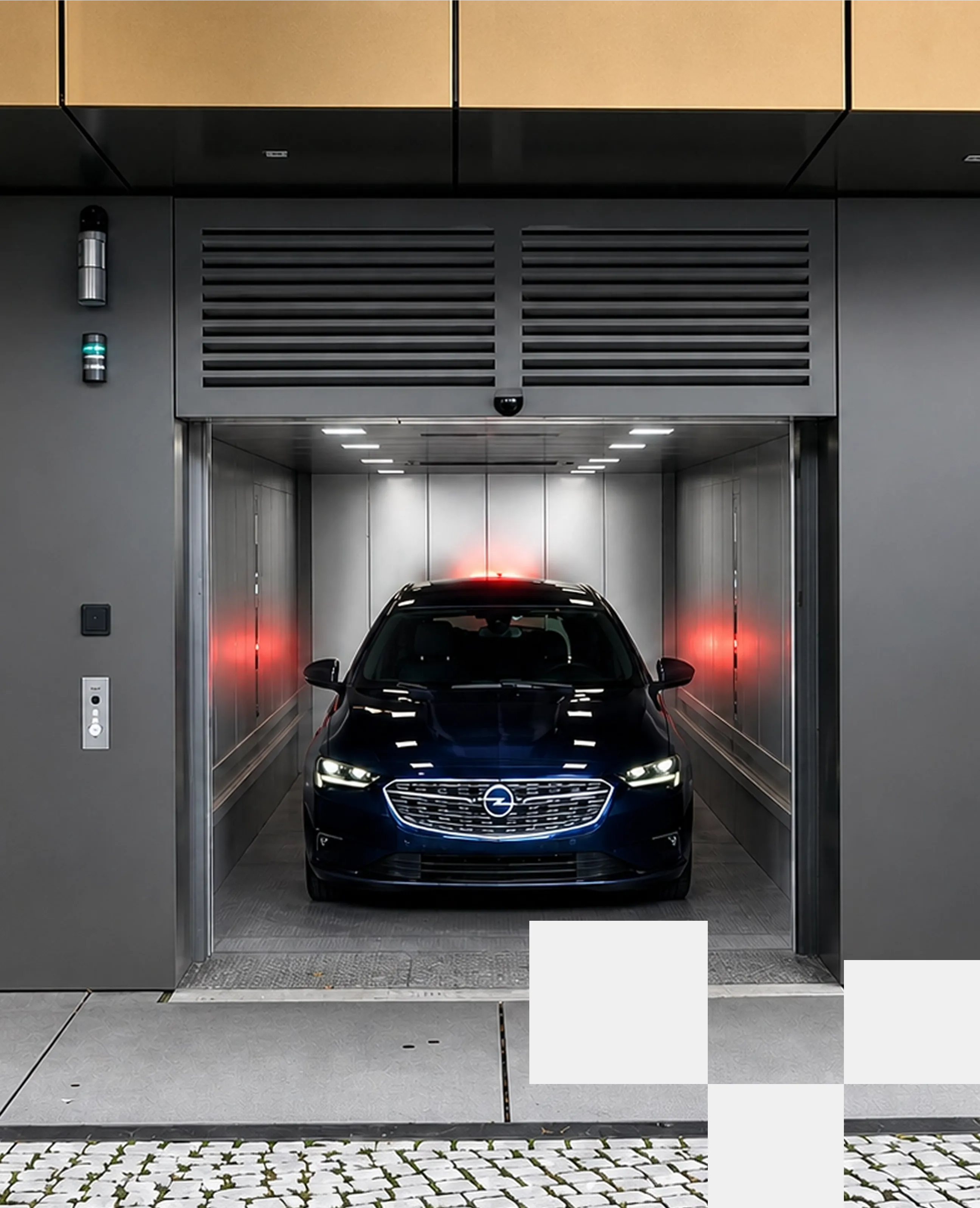Front view of a dark blue Opel car with headlights on, inside a modern car elevator with metal walls and ceiling lights.