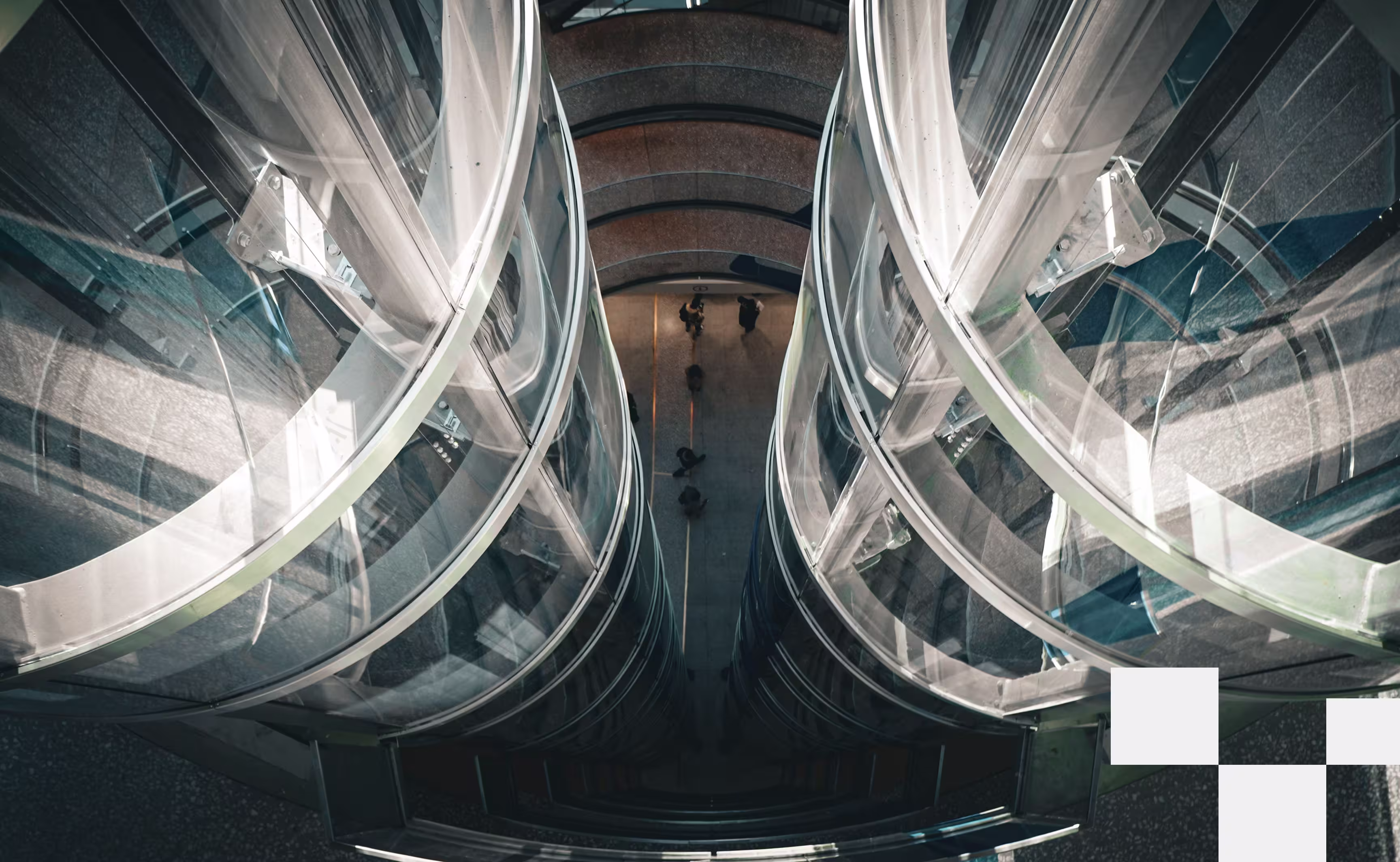 An image looking down into a busy walking intersection, flanked by 2 parallel glass tubes that contain elevators inside of them.