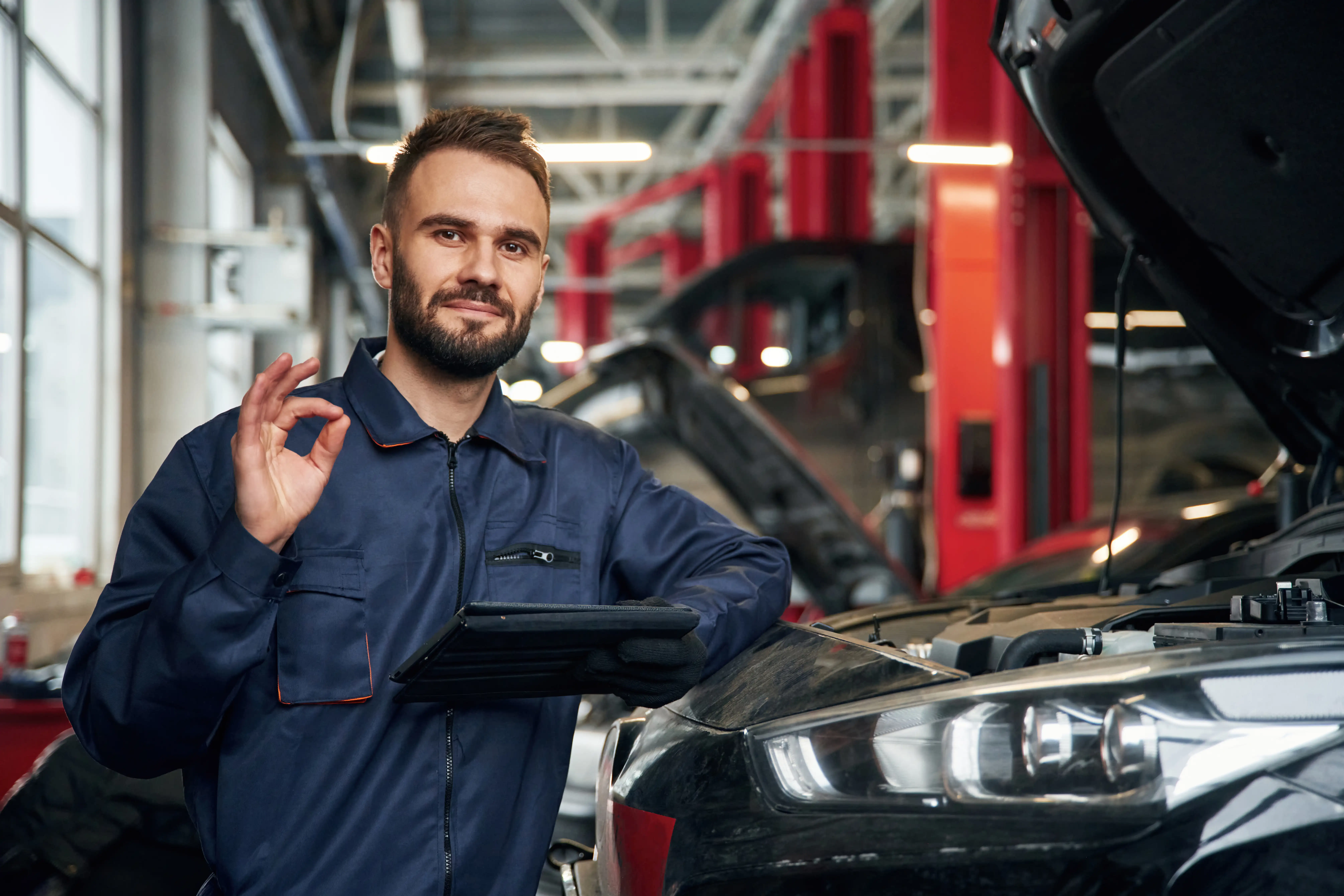 A mechanic in a blue jumpsuit gives an OK sign, smiling confidently next to an open car hood in a brightly lit garage, conveying professionalism.