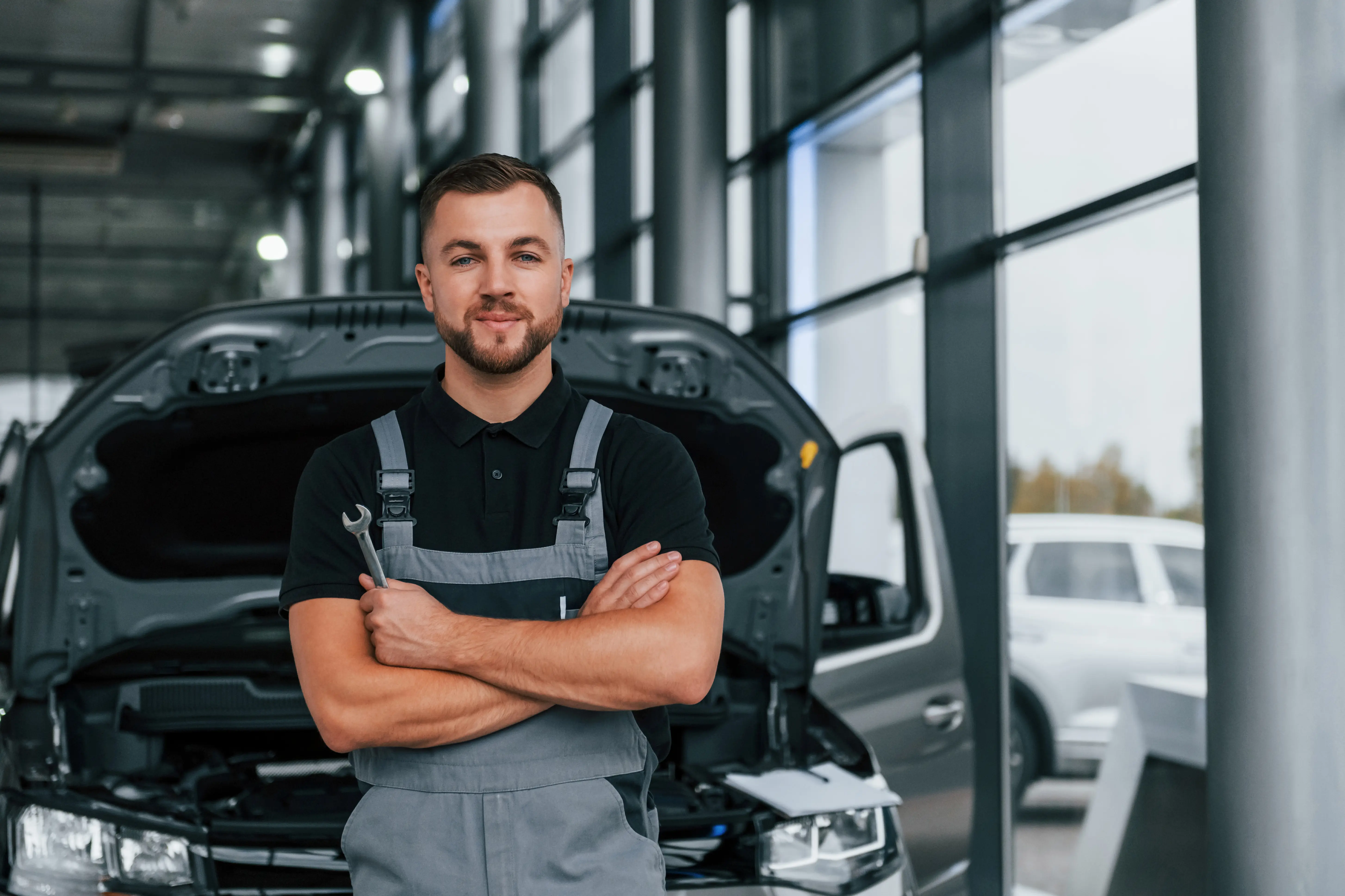 Mechanic in gray overalls stands confidently with arms crossed holding a wrench in an auto repair shop. Open car hood in background, bright windows beside.