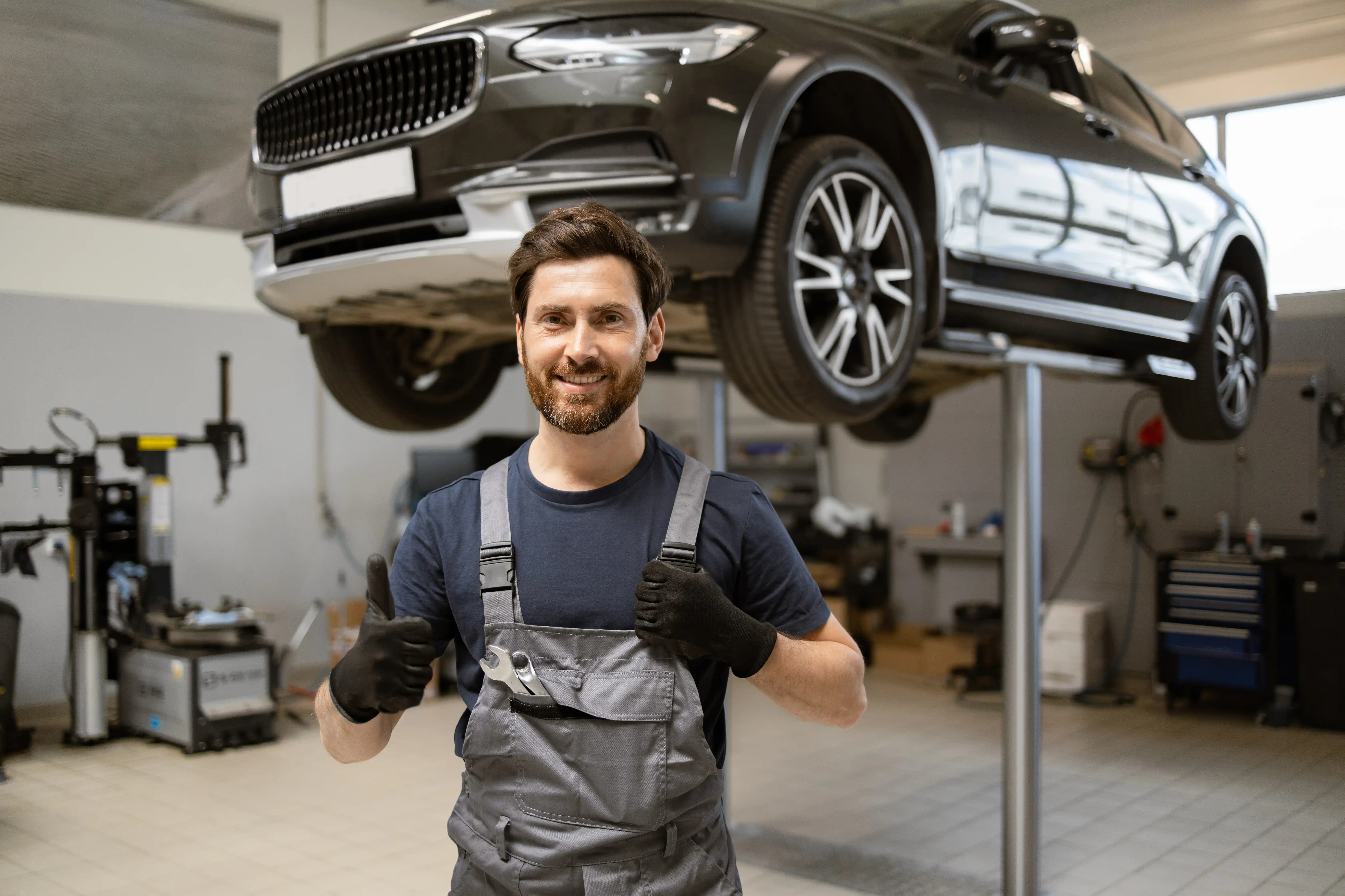 A smiling mechanic in a workshop stands confidently in front of a car lifted on a hoist, wearing gray overalls and black gloves. The scene conveys a sense of professionalism and readiness.