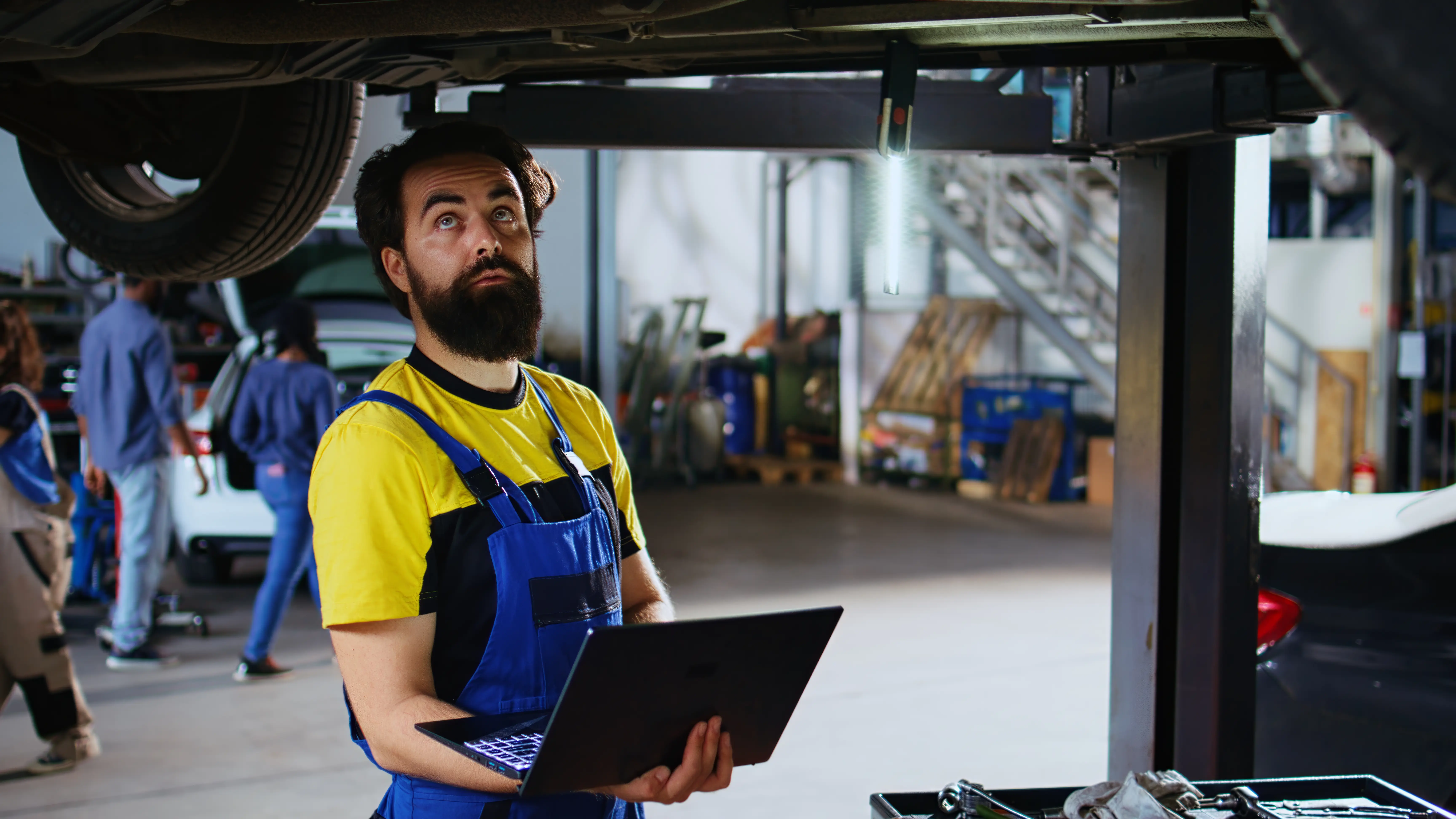 A mechanic in blue overalls intently examines a car's underside, holding a laptop. The workshop background is busy, with tools and vehicles visible