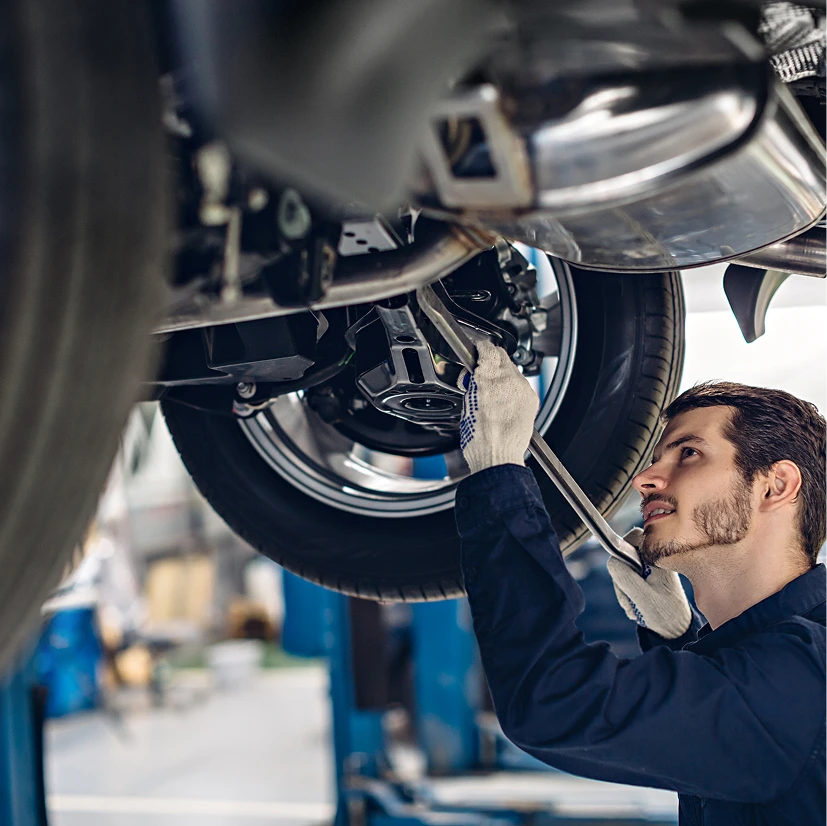 A mechanic wearing gloves and a blue jumpsuit works under a vehicle on a lift, using a wrench. He appears focused and the scene is well-lit.