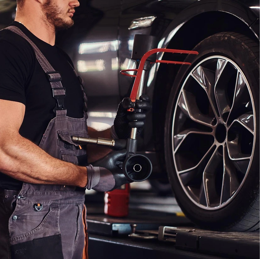 A mechanic in overalls and gloves holds a tool while preparing to work on a vehicle tire in a garage. The scene conveys focus and expertise.