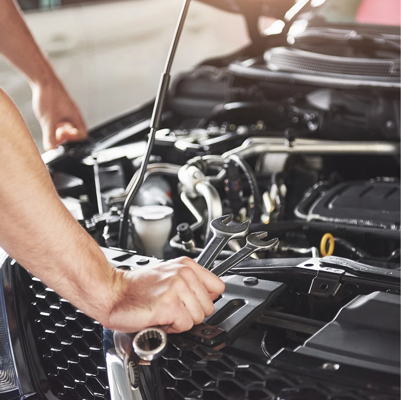 A mechanic's hands hold wrenches while inspecting a car's engine. The open hood and tools suggest ongoing maintenance in a well-lit garage.