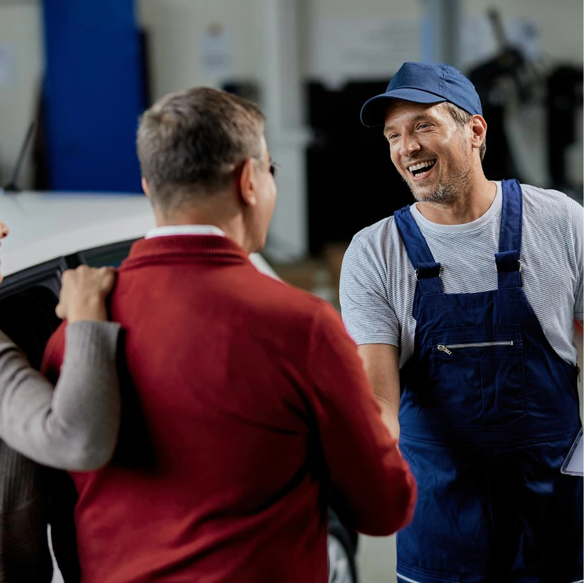 A mechanic in blue overalls and a cap warmly shakes hands with a smiling customer in a red sweater, conveying a friendly and positive interaction in an automotive workshop.