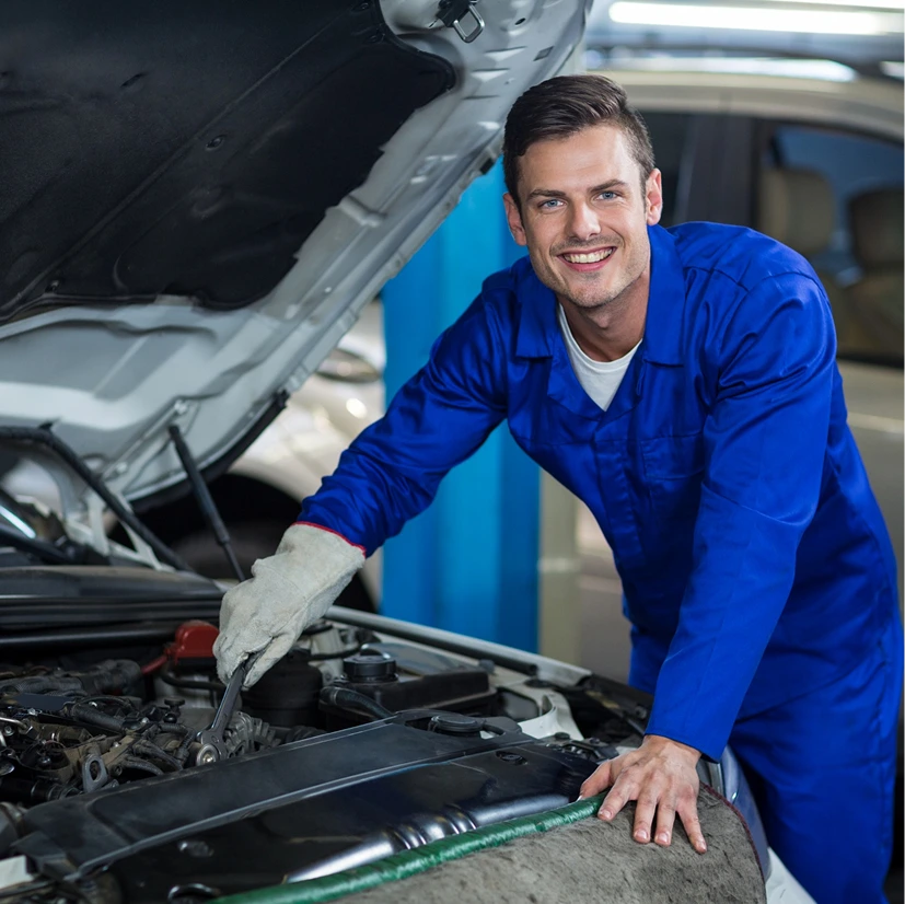 Smiling mechanic in blue overalls working under an open car hood, holding a wrench. The scene is in a bright, organized garage, conveying a positive, professional atmosphere.
