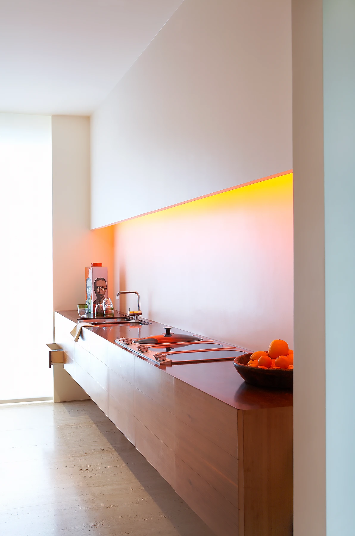 The kitchen with a floating drawer unit and worktop to one side and floor-to-ceiling storage to the opposing side, lit by colour-changing light washing the wall and ceiling.