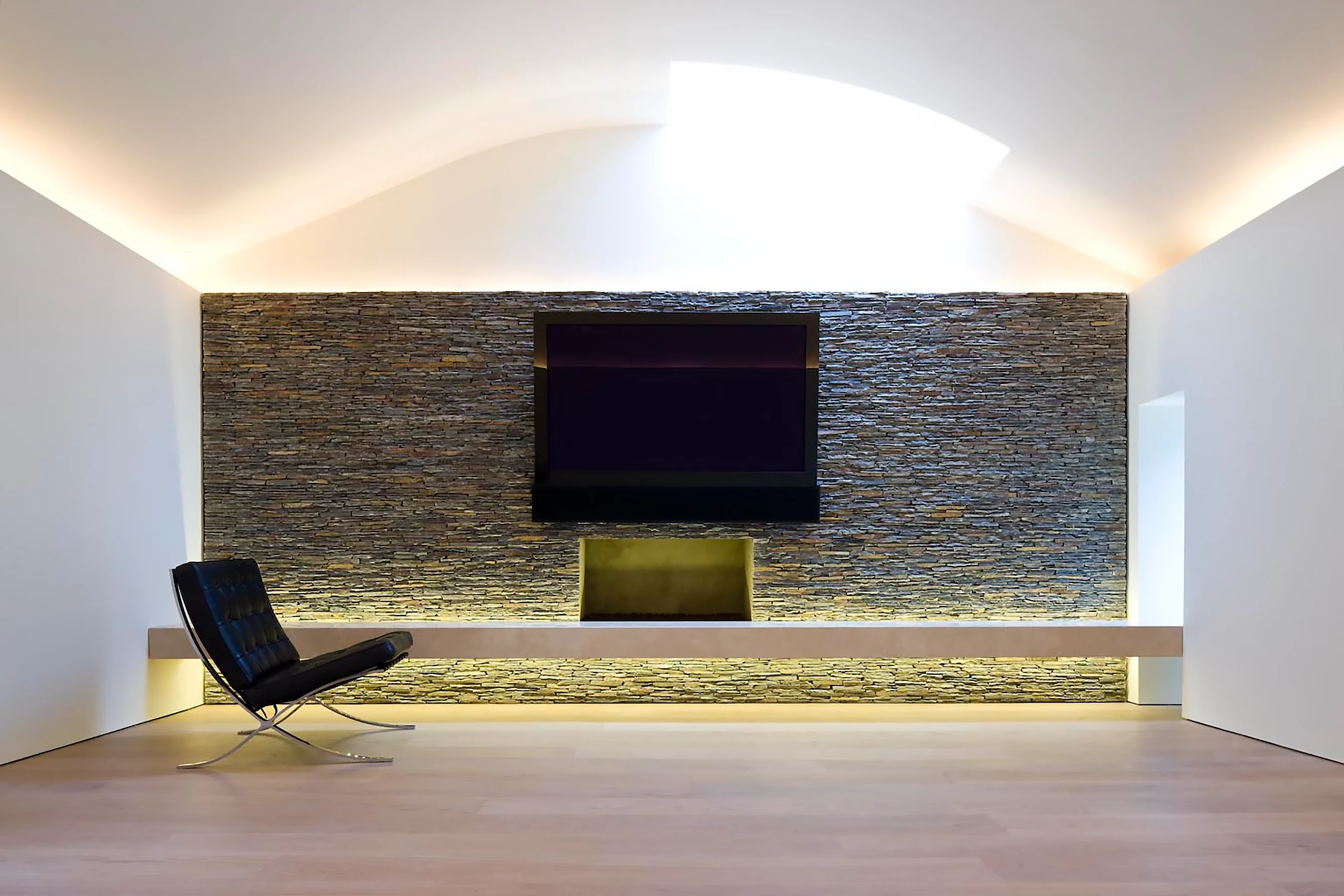 The first-floor gallery with an Andy Goldsworthy-style hand-built slate wall. The wall is washed with light from behind the travertine cantilevered bench. The bench itself appears weightless as it floats away from the wall and projects through the glass to the garden beyond. 
