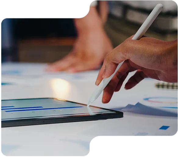 A hand uses a stylus on a tablet displaying charts on a desk with papers and blurred background.