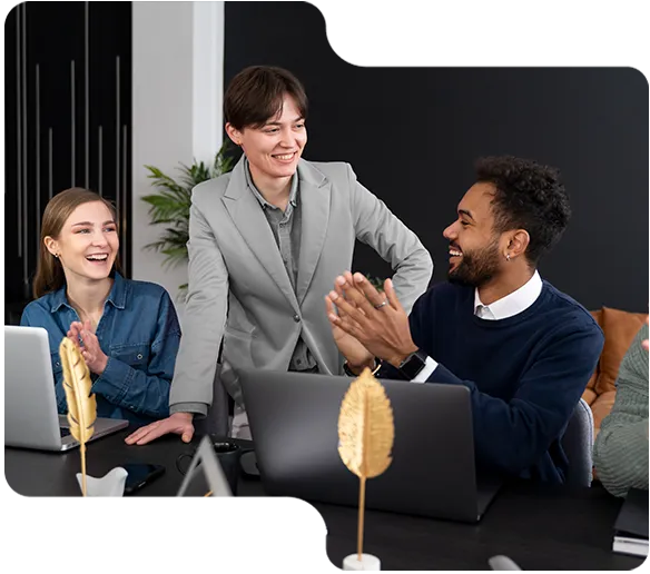 Three colleagues smiling and applauding in a modern office with laptops on the table.
