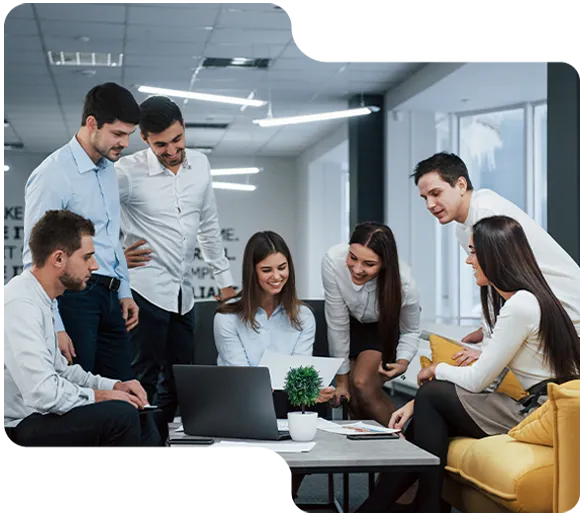 Seven professionals gathered around a laptop and papers in a modern office, engaged in discussion.