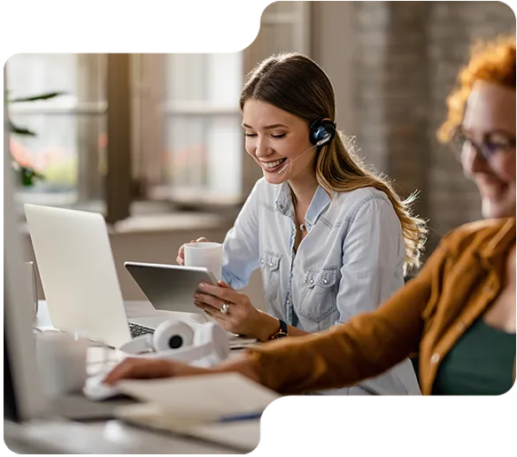Smiling woman with headset using tablet and laptop at bright office with coworker.