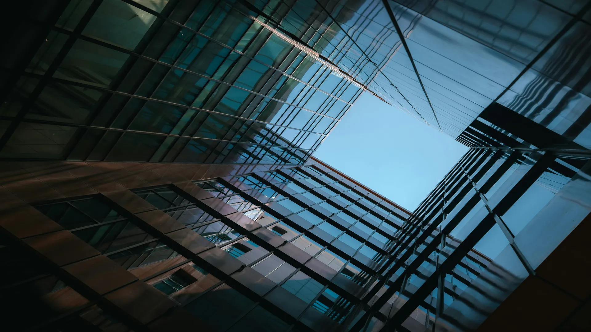 Low-angle view looking up between modern glass office buildings, with reflective blue-tinted windows and intersecting architectural lines framing a bright blue sky overhead.