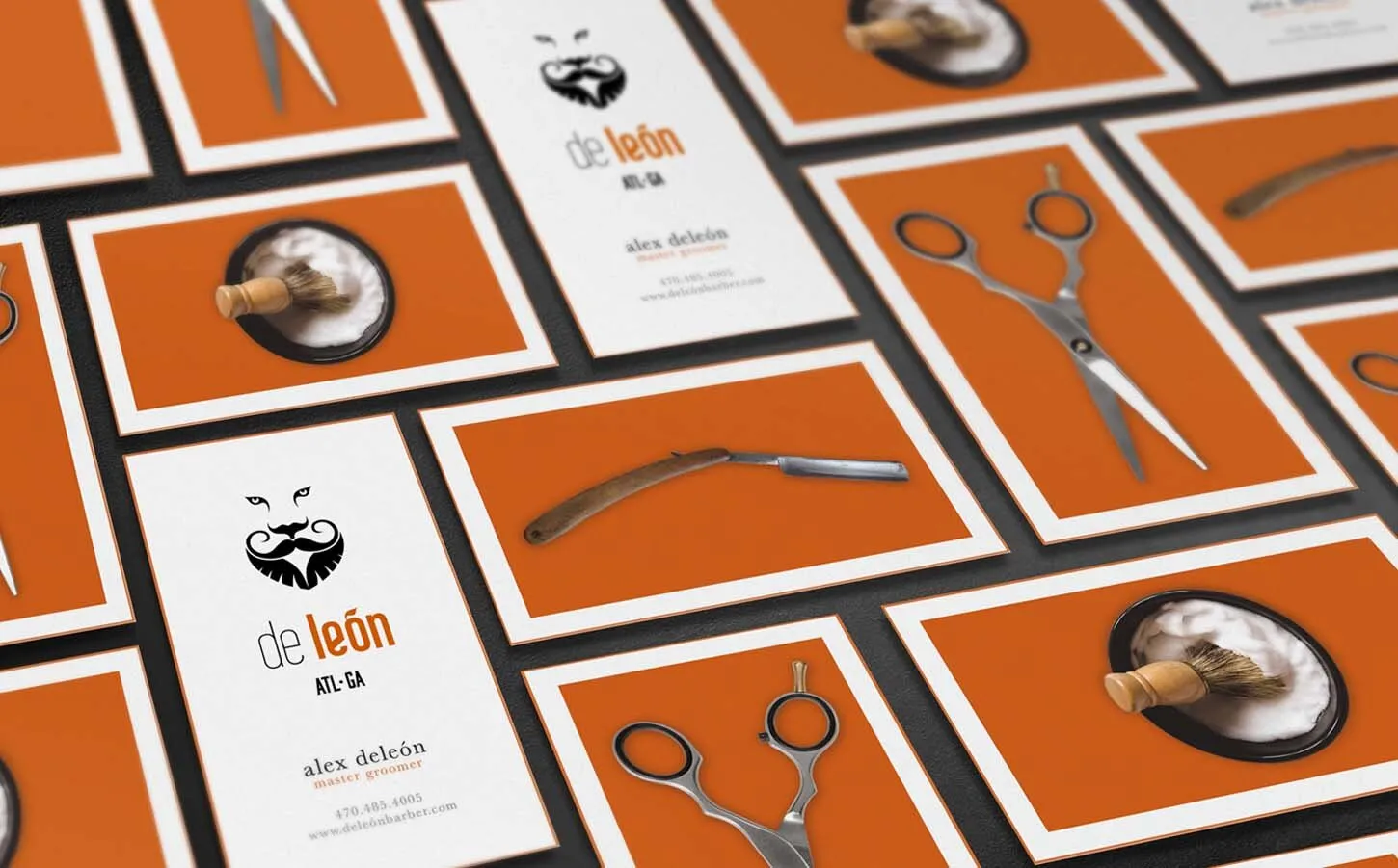 De León business cards spread across a surface, orange cards carrying the lion and leaf mark alongside dark cards and a pair of barber shears