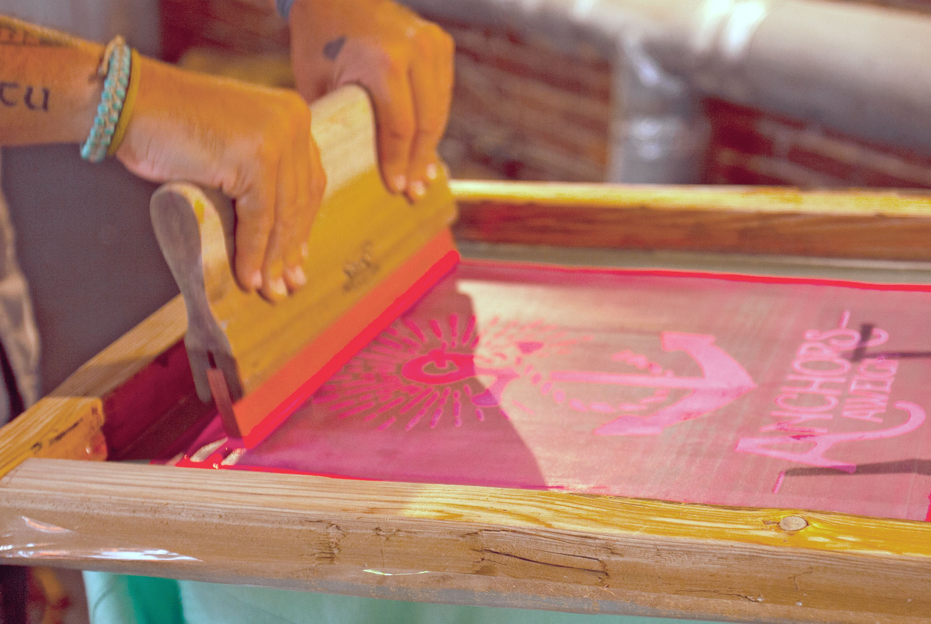Hands pulling a squeegee across a J. James screen, printing an anchor and the words Anchors Aweigh in pink ink