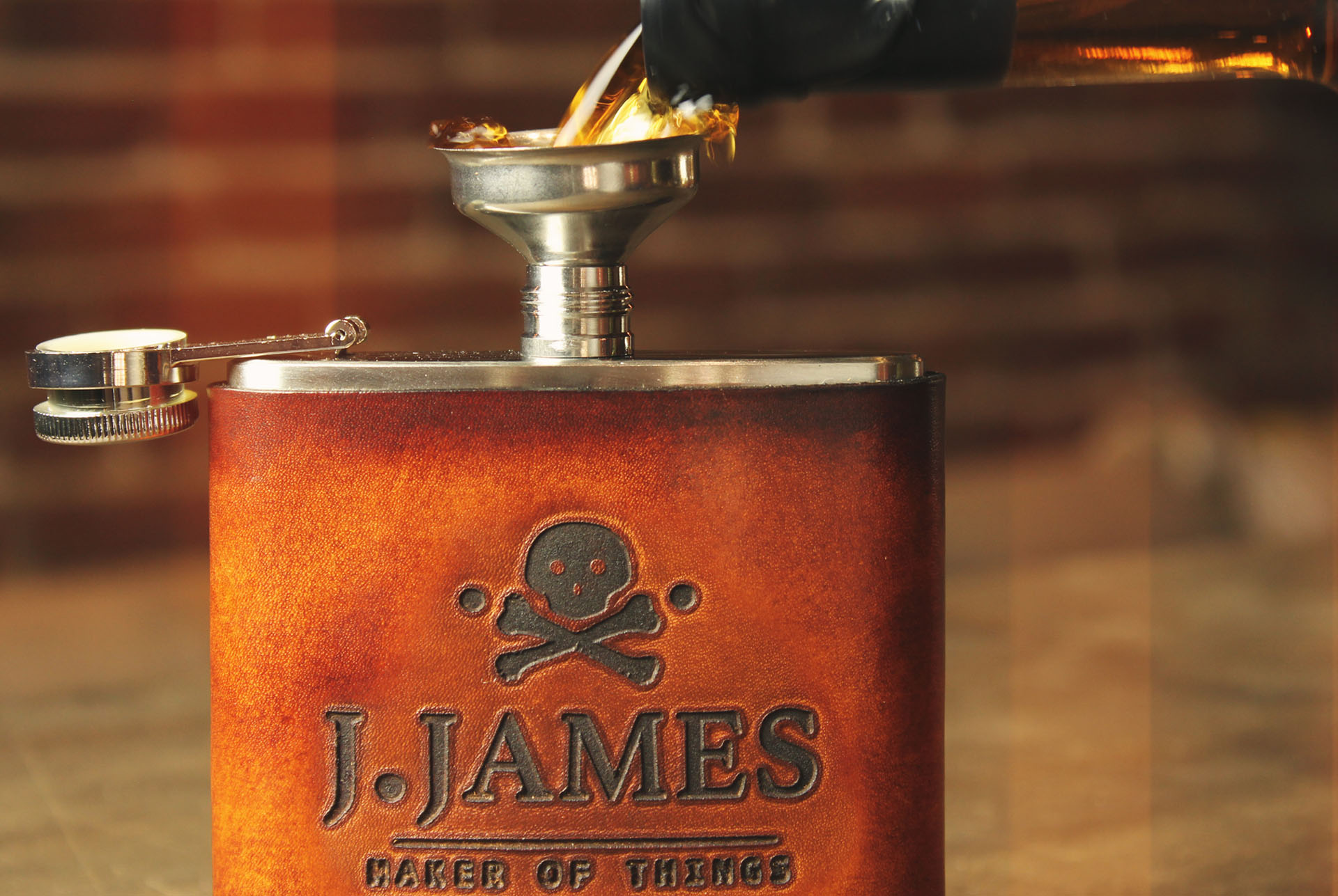 A J. James leather wrapped flask being filled through a funnel, the tooled wrap carrying the skull mark above J. JAMES MAKER OF THINGS