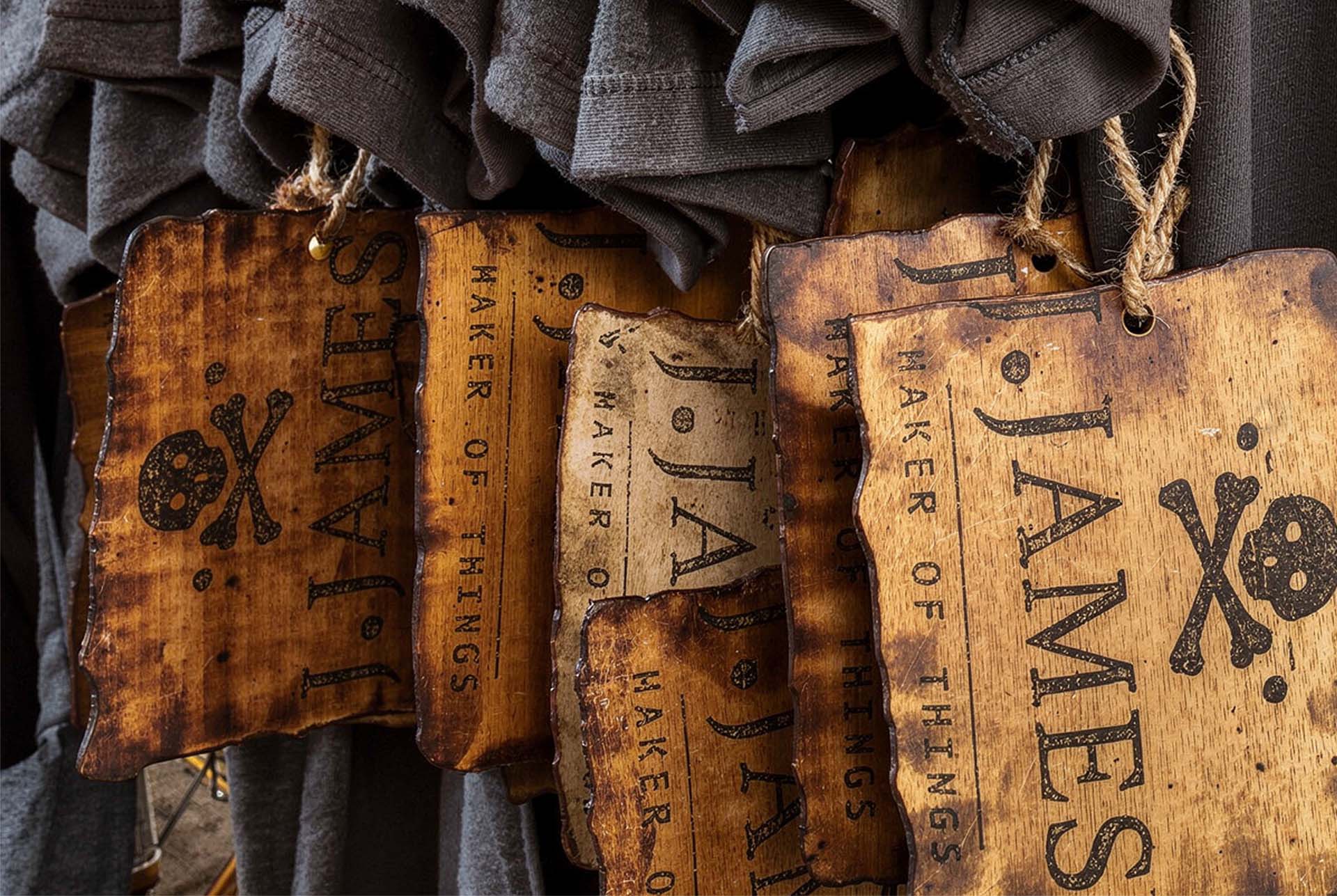 A row of J. James apparel hangtags hanging on cords, each with the skull mark and wordmark burnt into the wood