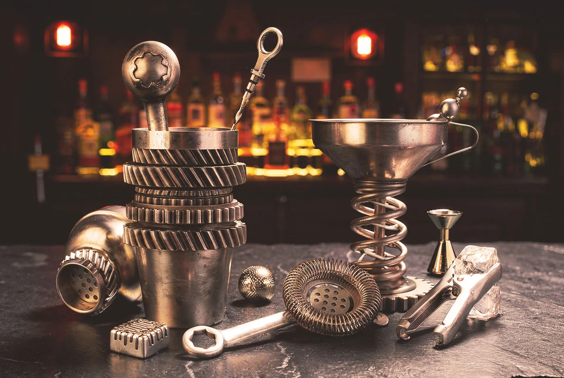 Gearbox bar tools on a stone bar top with a lit back bar behind, a gear body shaker on its side, a coil spring stemmed funnel, a hawthorne strainer, a jigger and a corkscrew