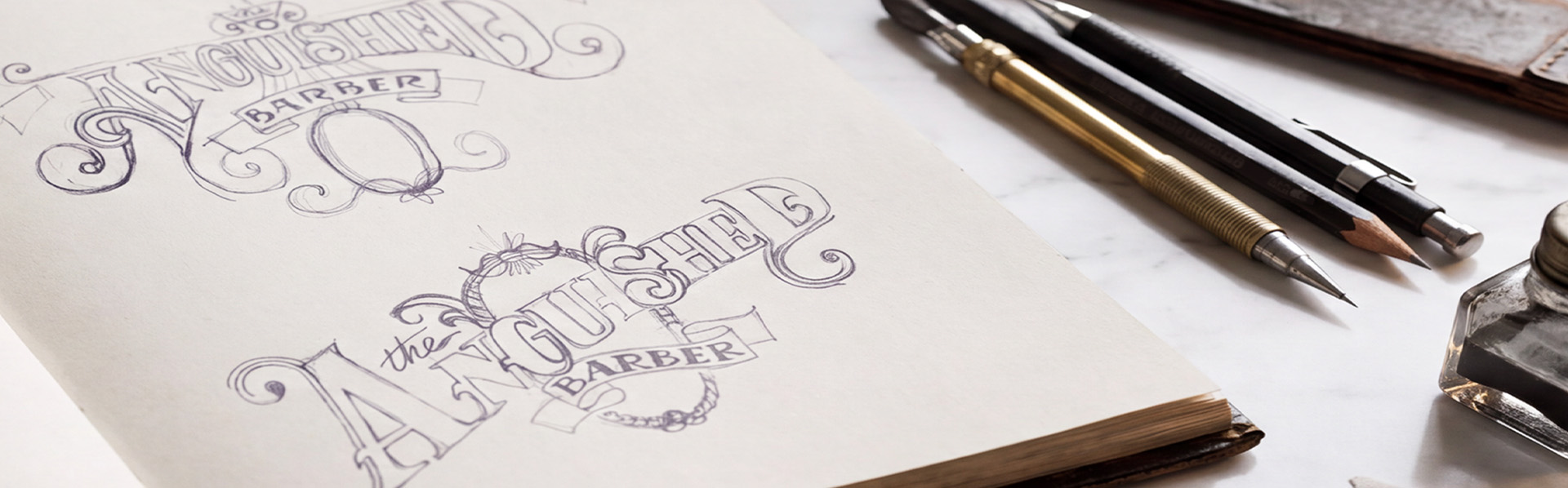 Hand drawn ornate lettering sketches for The Anguished Barber logotype in a sketchbook, beside pencils, dip pens and ink