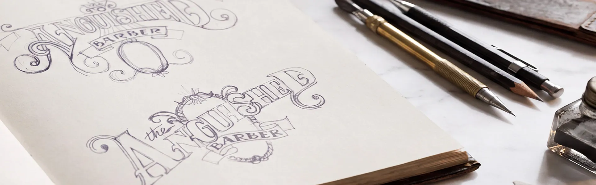 Hand drawn ornate lettering sketches for The Anguished Barber logotype in a sketchbook, beside pencils, dip pens and ink