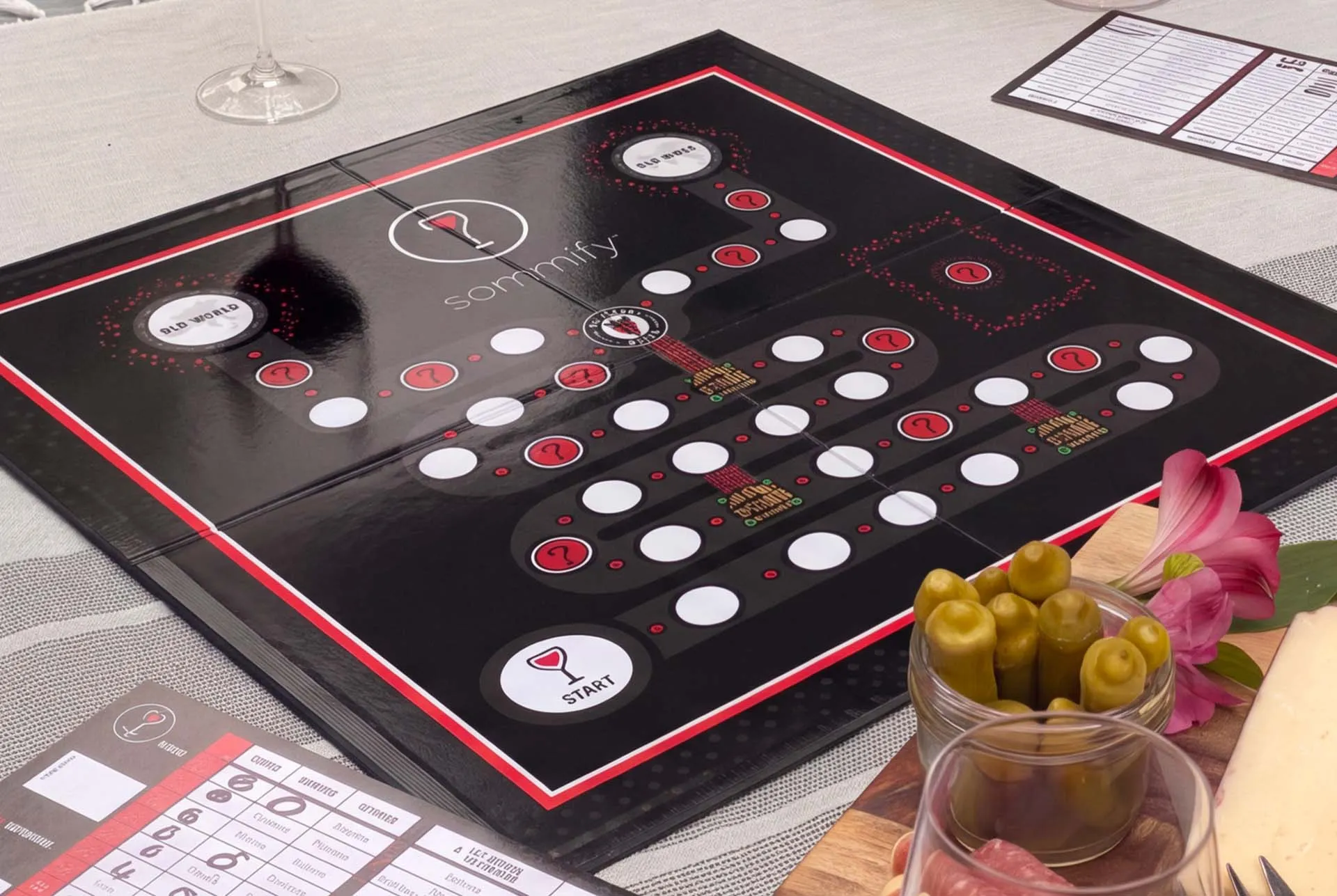 The printed Sommify board open on a table mid game, score sheets to one side and a bowl of olives and flowers alongside