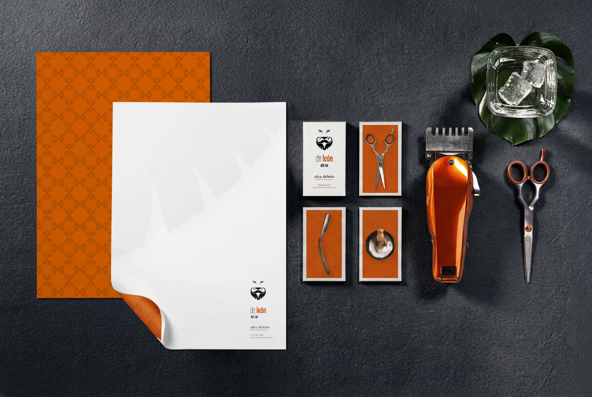 De León stationery flat lay on dark slate with letterhead, business cards, an orange comb, barber shears and a Monstera leaf